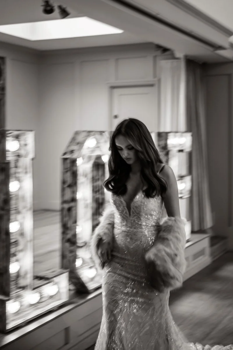 A woman in a sparkly gown with a fur stole standing in front of vanity mirrors with lights in a room.