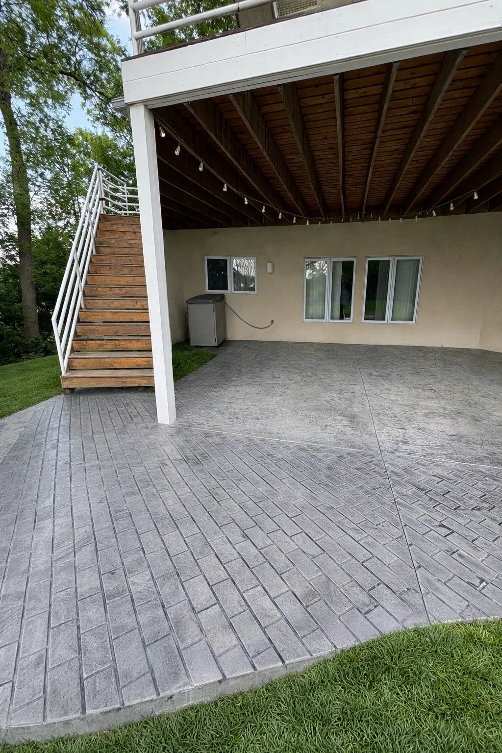 A covered patio with a concrete and brick-patterned floor, a beige wall with three windows, a washer, and string lights hanging from the ceiling. An exterior staircase with wooden steps and white metal railings leads to the upper level. Trees and grass are visible around the patio.