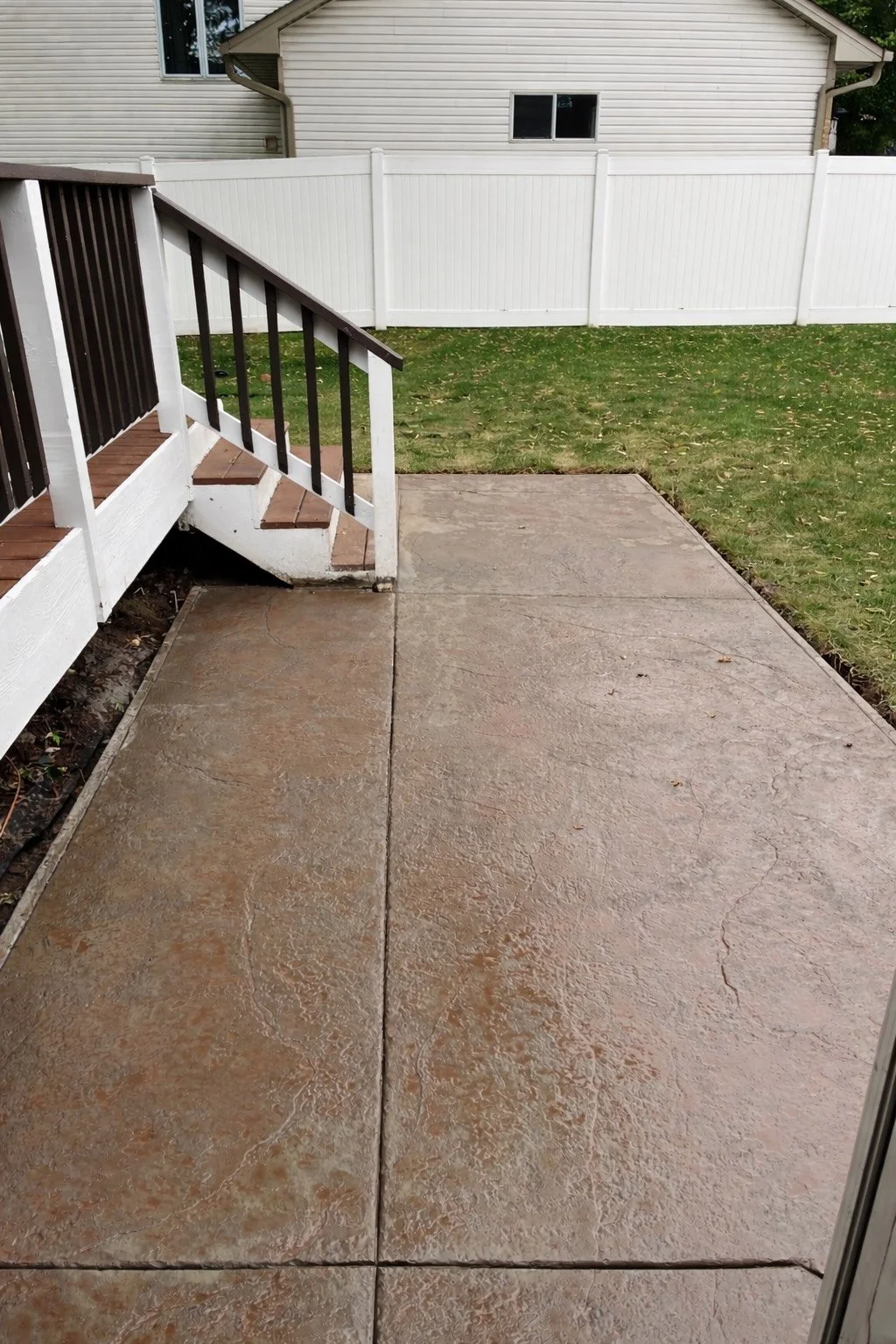 Backyard patio with concrete surface, white railing stairs leading down to grass, white vinyl fence, and a backyard with a house in the background.