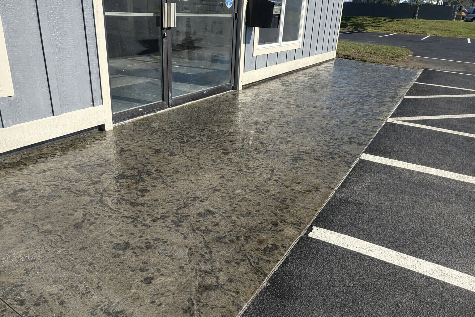 Outside of a building with a concrete sidewalk and a parking lot. The building has a glass door and gray wooden siding, and the sidewalk appears to be wet.