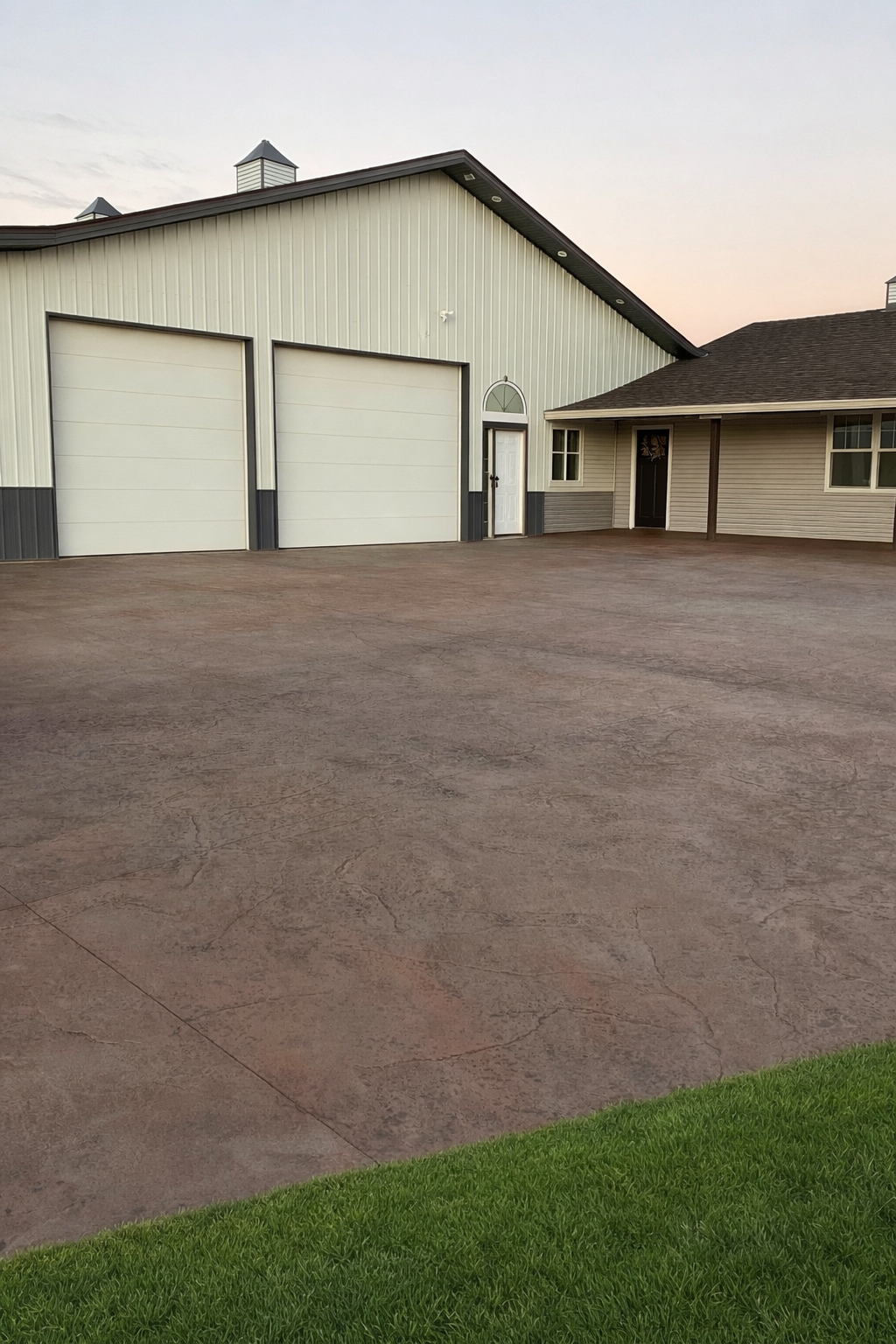 A large concrete driveway in front of a house with a two-car garage, a door, and windows. The house has beige siding and a dark-colored roof, with a patch of green grass in the foreground.