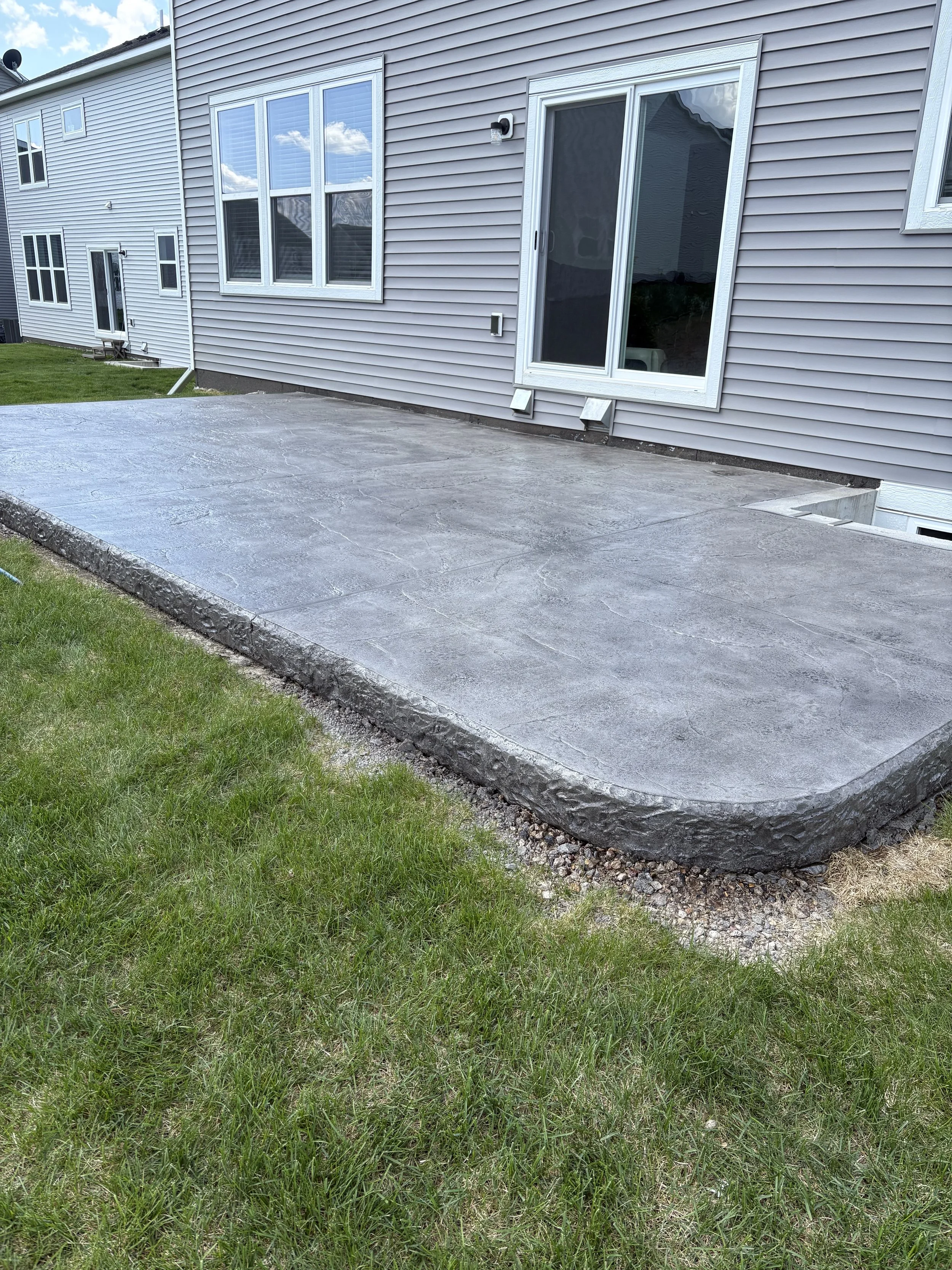 Newly poured concrete patio in backyard with sliding glass door and windows on house, green grass surrounding the patio, and neighboring house visible in the background.