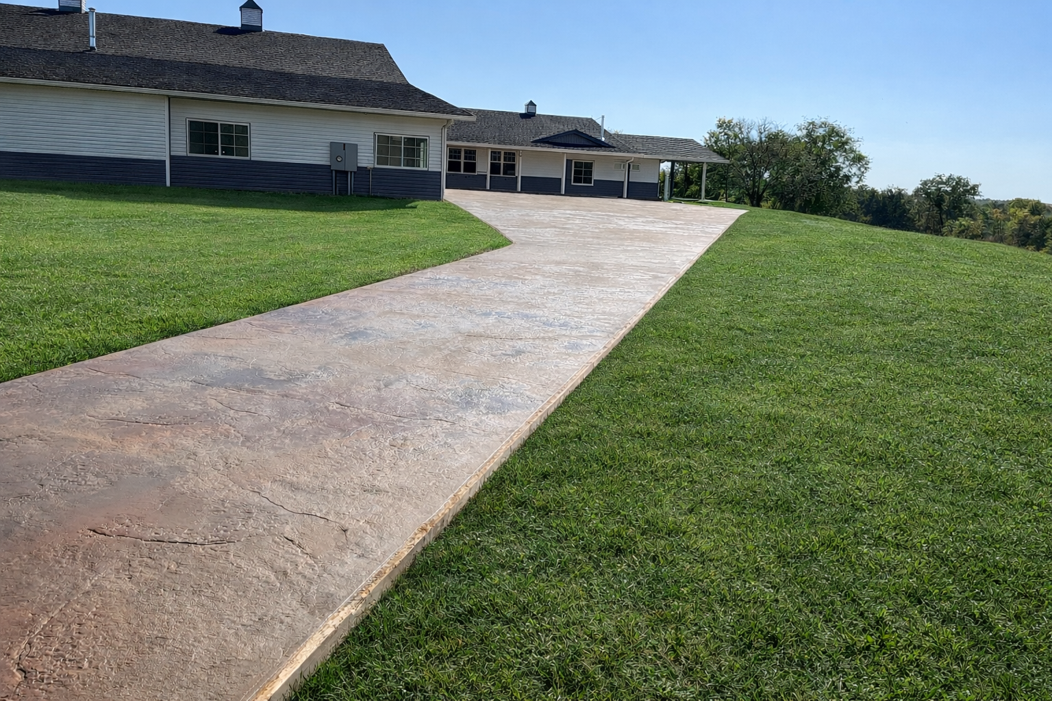 Concrete pathway leading up to a house with a porch, surrounded by green grass and trees, under a clear blue sky.