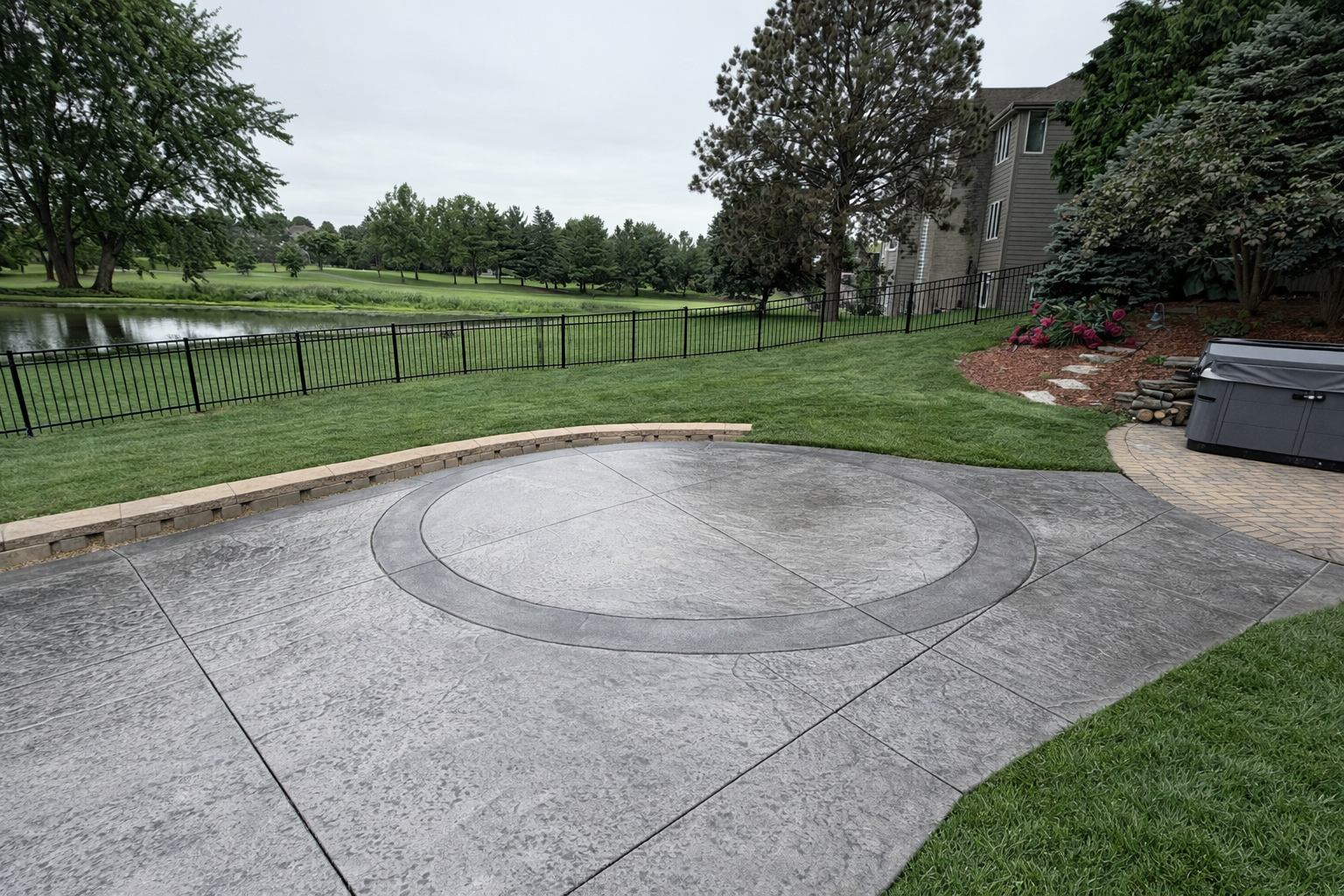 Backyard patio with stamped concrete, a raised garden bed with flowers, a black metal fence, a pond and trees in the background.