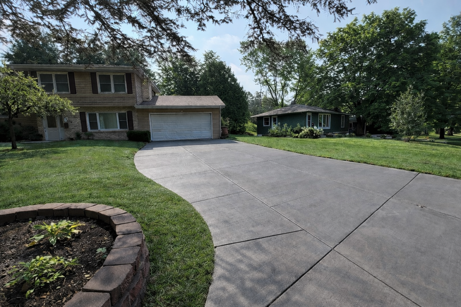 A curved concrete driveway leading to a two-story house with beige siding and brown shutters on the left, and a small green house with white trim on the right, surrounded by trees and green lawns under a partly cloudy sky.
