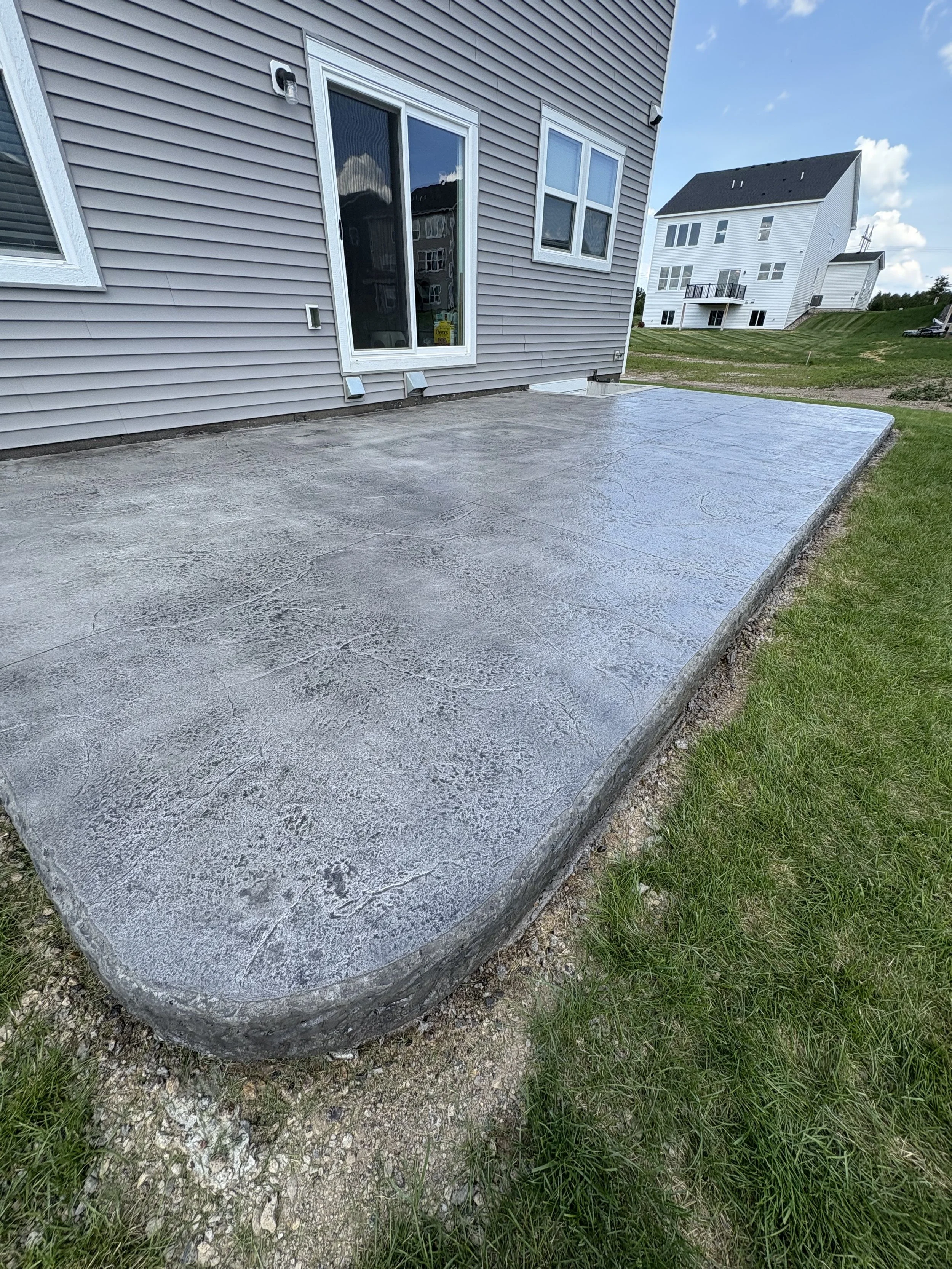 Freshly poured concrete patio outside a house with gray siding, sliding glass door, and windows, in a suburban neighborhood on a sunny day.