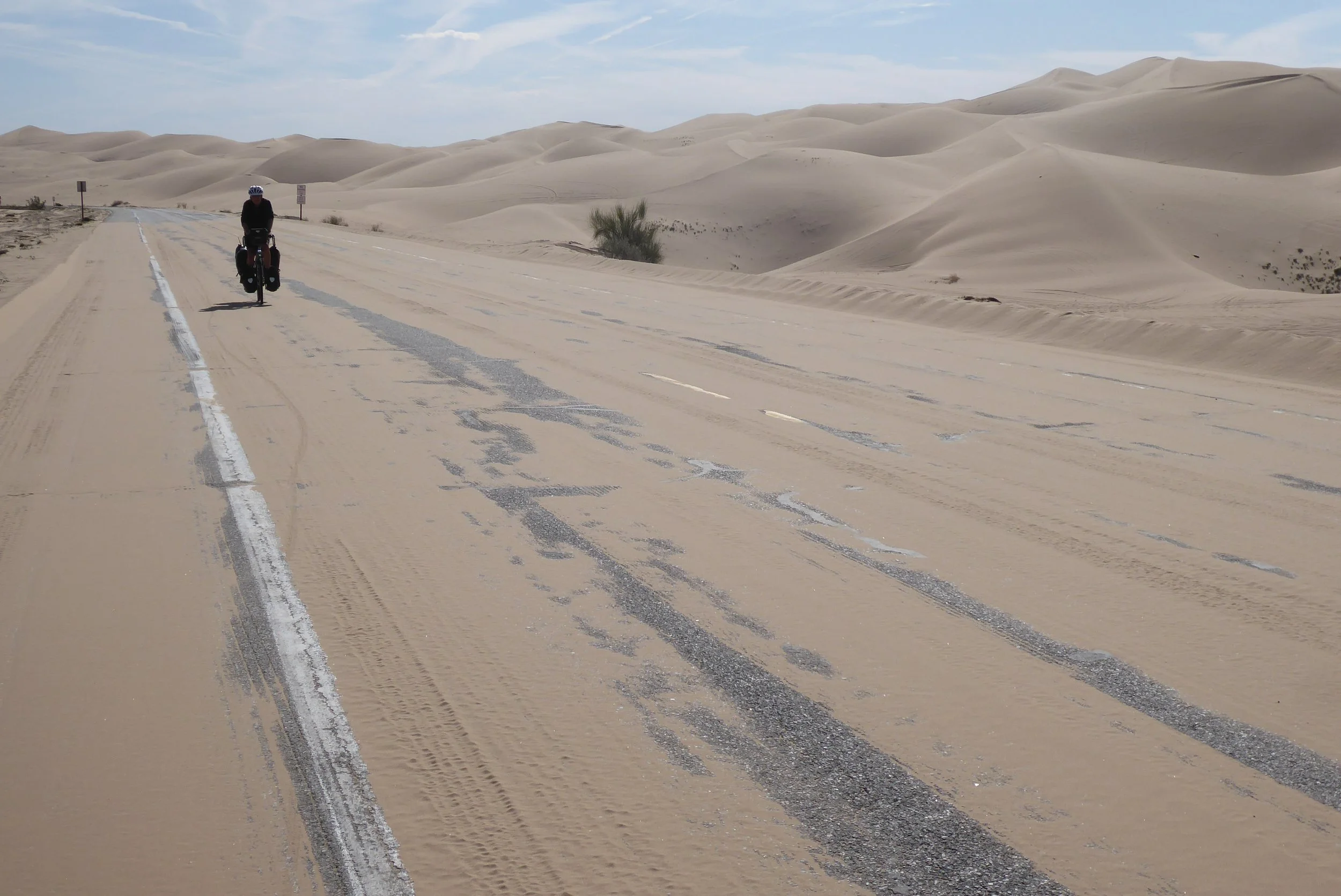 Person riding a bicycle on a paved road through a desert with sand dunes under a partly cloudy sky.