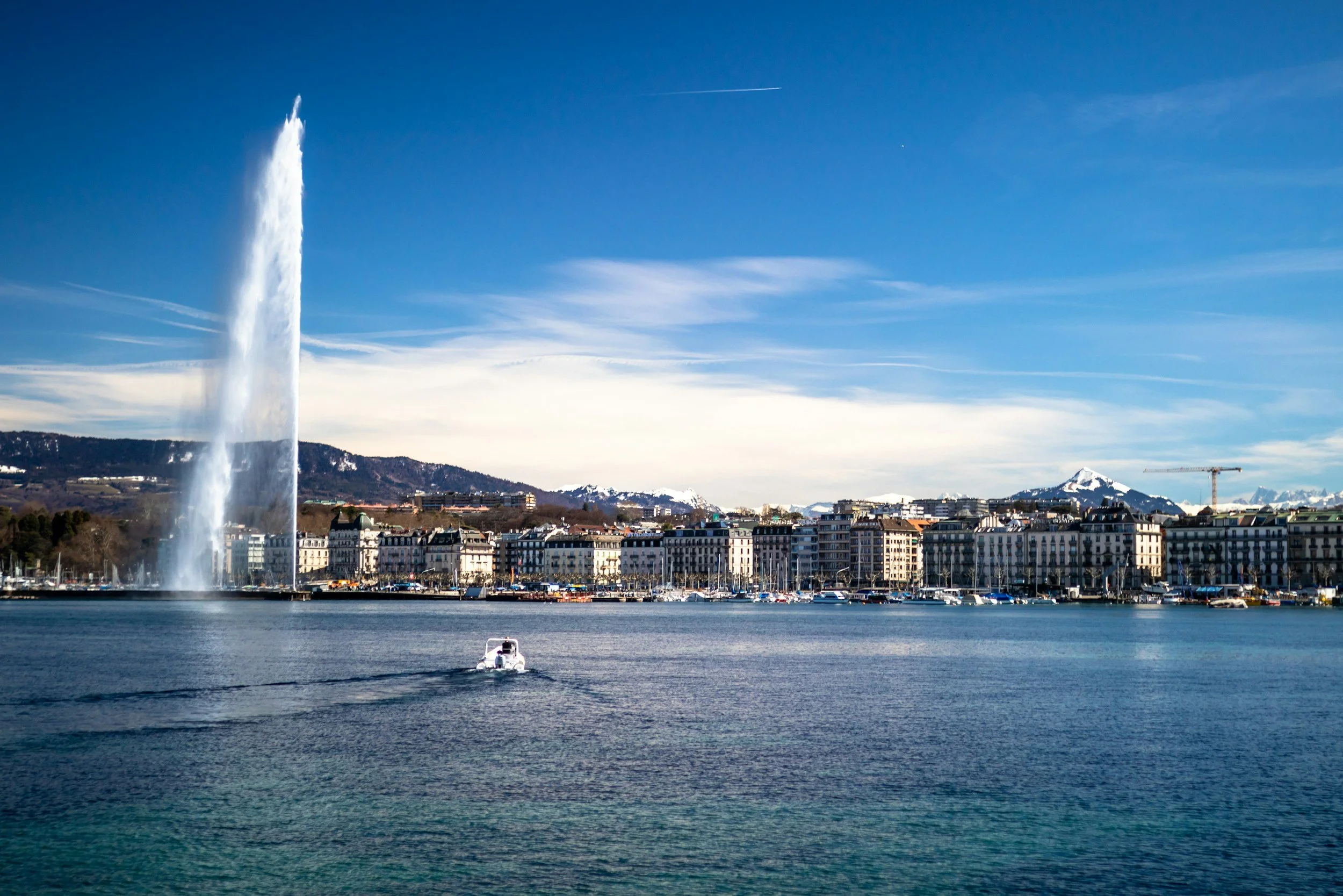 A large fountain shooting water high into the sky on a lake, with a cityscape along the shore, mountains in the background, and a boat floating on the water in the foreground.