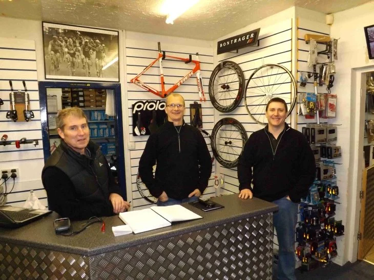 Three men standing behind a counter inside a bicycle shop with bike frames, wheels, and accessories on display.