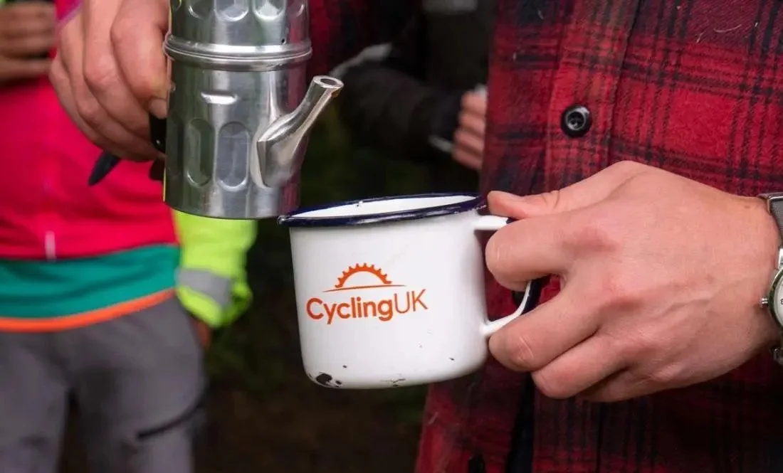 Person holding a white Camping UK enamel mug with a spout, while pouring hot water from a small metallic kettle. The person is wearing a red plaid shirt and a wristwatch.