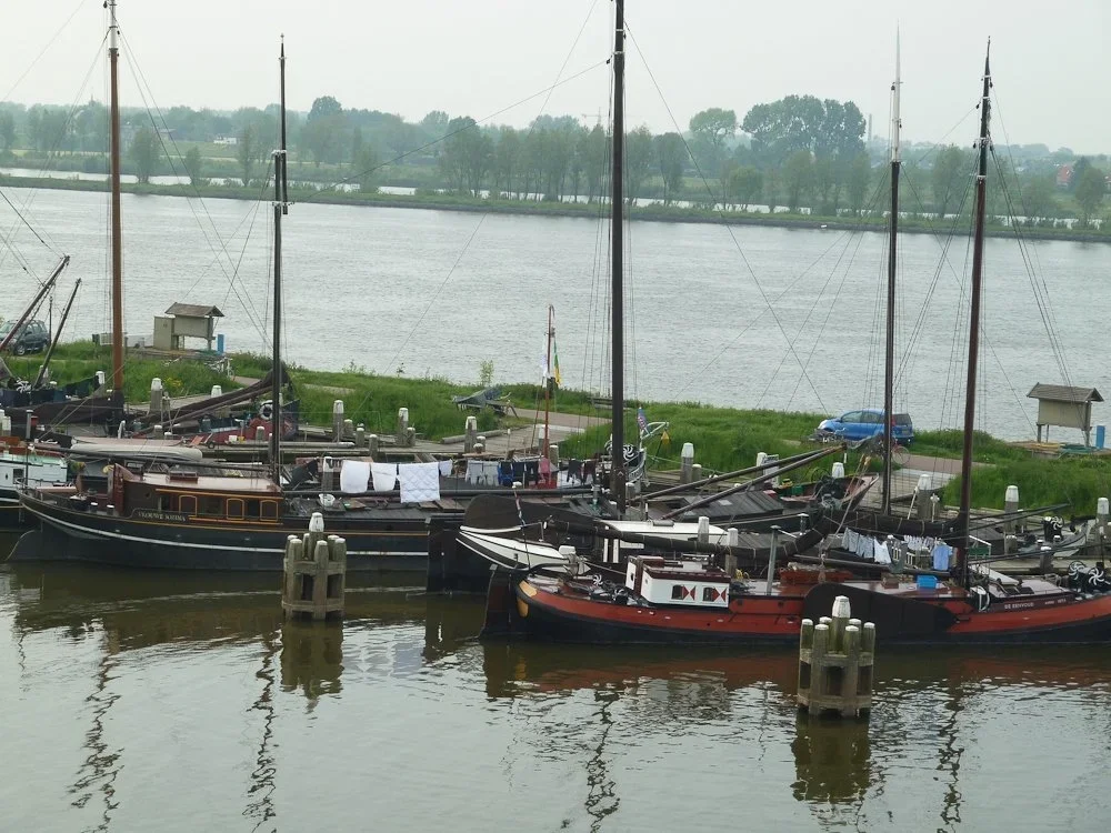 Several sailboats docked at a marina along a river, with clothes hanging on a line and a grassy area in the background.