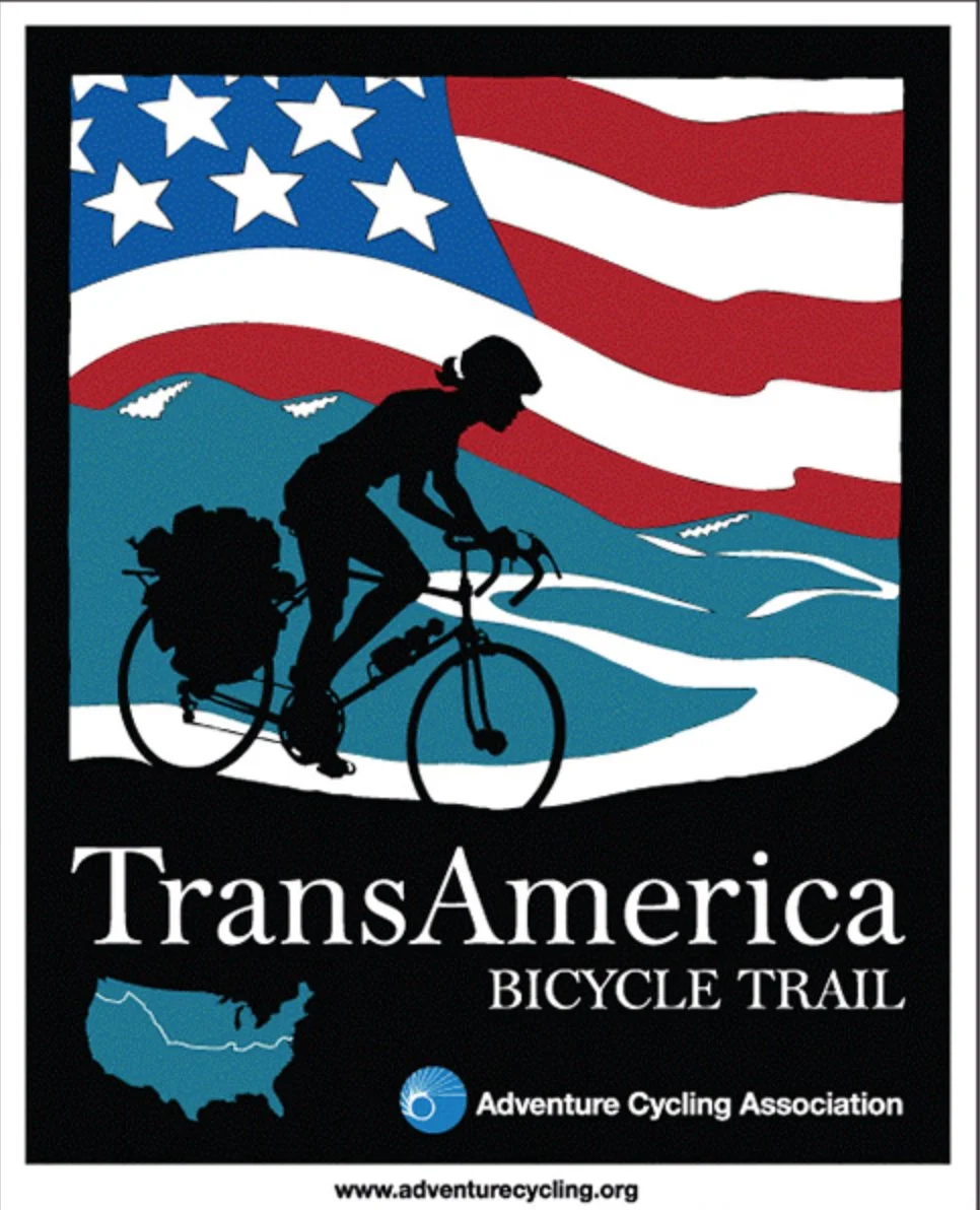 A poster for the TransAmerica Bicycle Trail featuring a cyclist silhouette with a large pack on a mountain bike set against an American flag background, with a map of the United States and a logo for the Adventure Cycling Association.