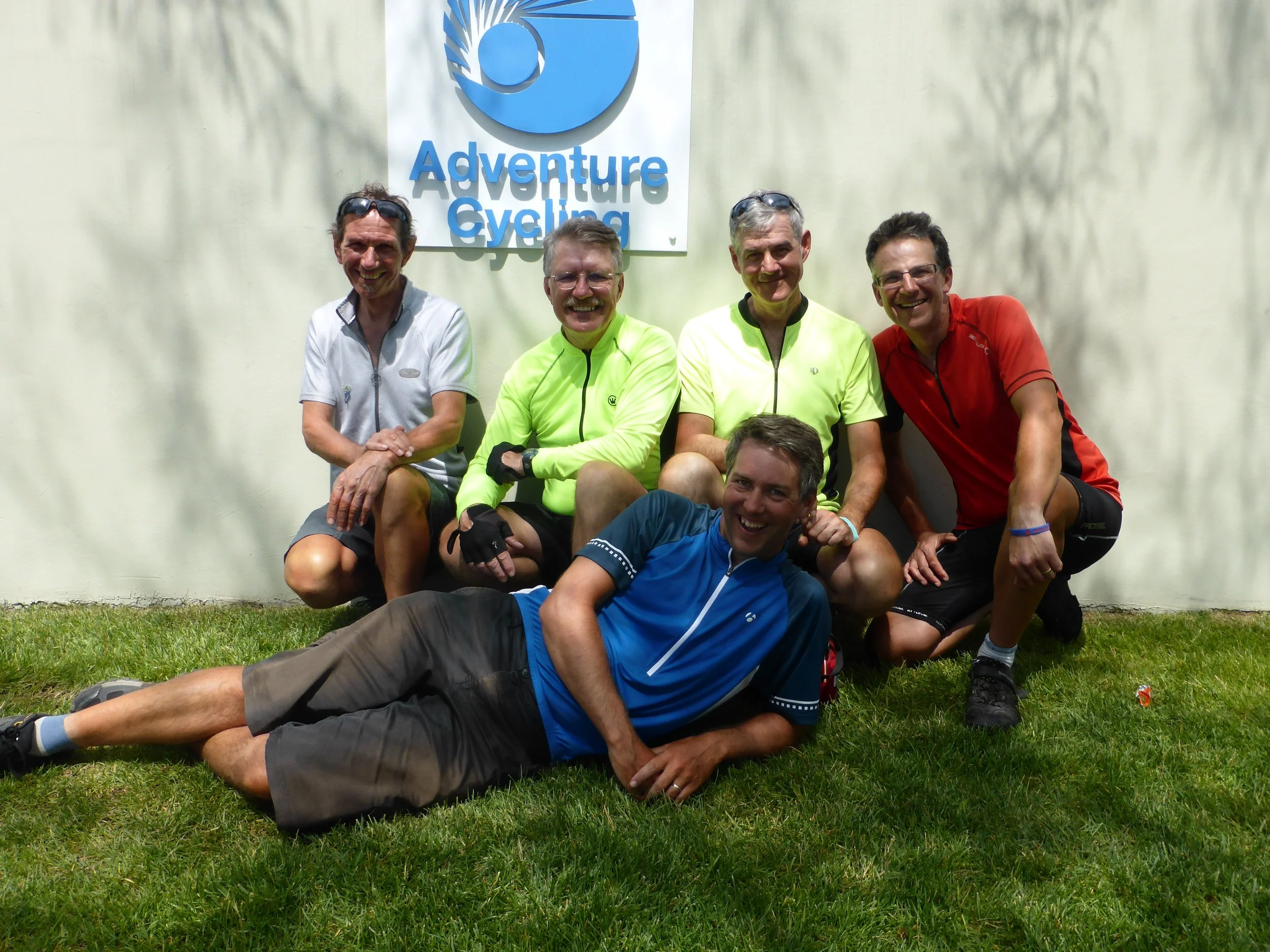 A group of five men posing for a photo in front of a sign that reads 'Adventure Cycling.' Four of the men are standing, and one is lying on the grass in front. They are all wearing cycling apparel and sunglasses, smiling happily.