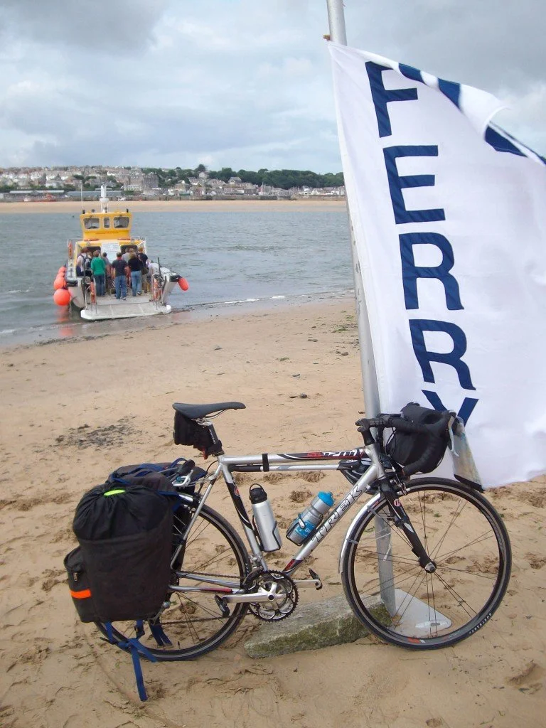 A bicycle with a black bag, water bottles, and a small pouch is parked on a sandy beach next to a white flag with blue writing that says "FERRBY". In the background, a yellow boat with a group of people on it is docked at the shore, with a town and houses visible across the water.