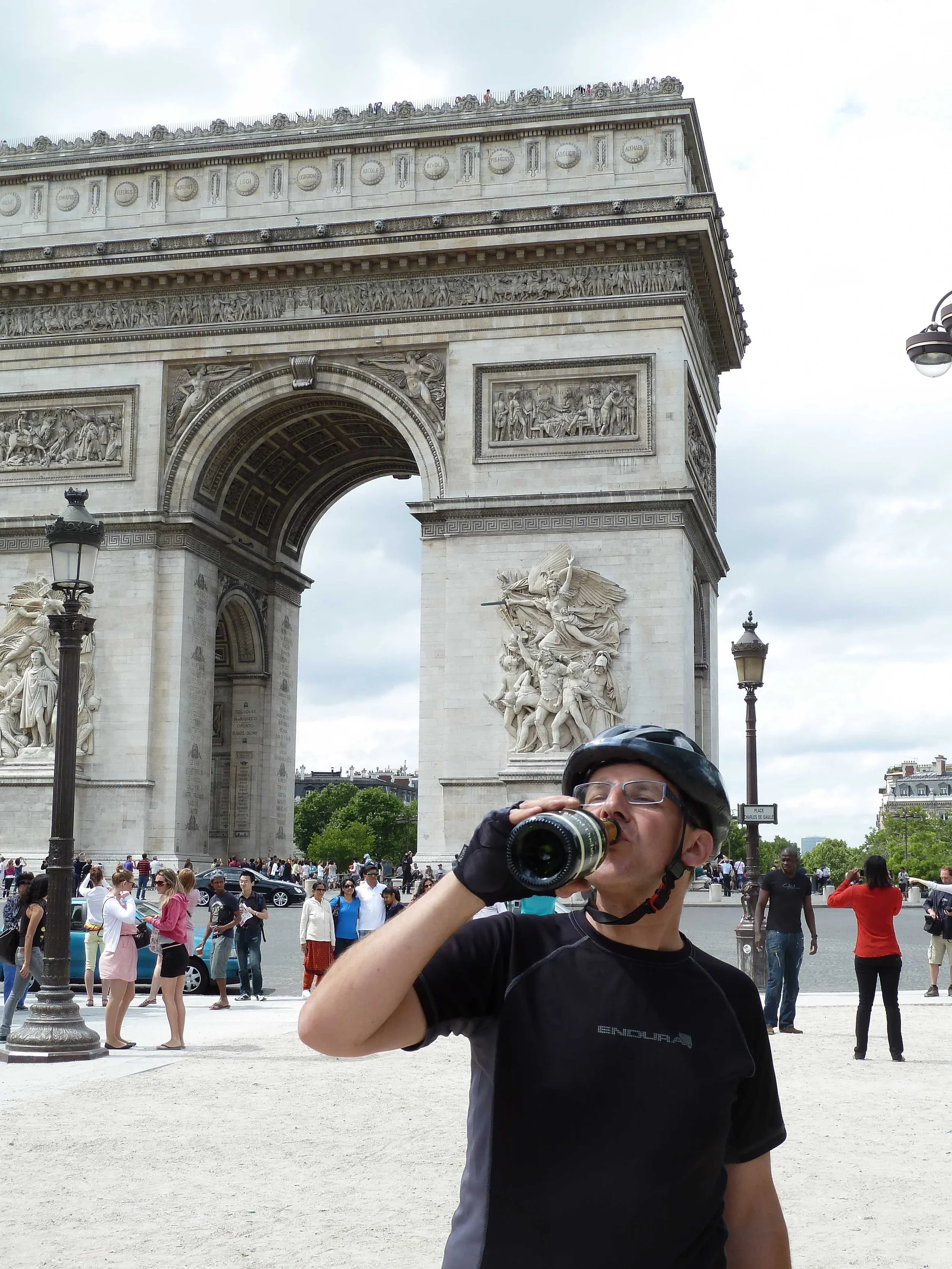 A man wearing glasses, a black helmet, and a black T-shirt is drinking from a bottle in front of the Arc de Triomphe in Paris, France. There are many people walking and taking pictures around the monument on a cloudy day.