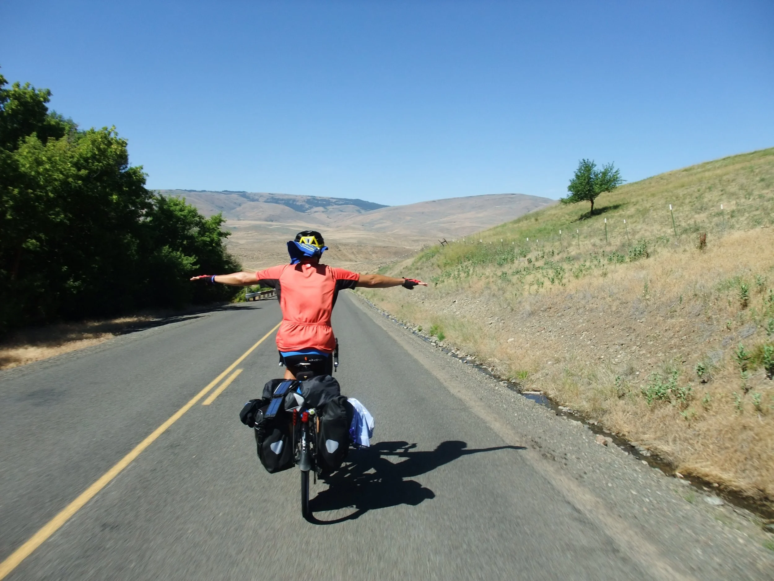 A person riding a bicycle on a country road with hills and trees in the background, arms outstretched.