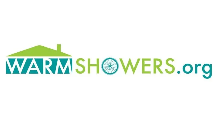 Logo for WarmShowers.org featuring a green house roof, the word 'WARM' in teal with a house chimney, and a bicycle wheel replacing the 'O' in 'SHOWERS' with the website name.