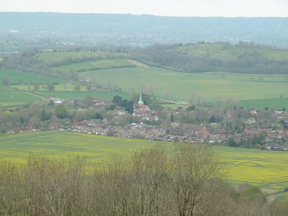 A panoramic view of a rural countryside with a small town, green fields, rolling hills, and a church with a tall steeple.