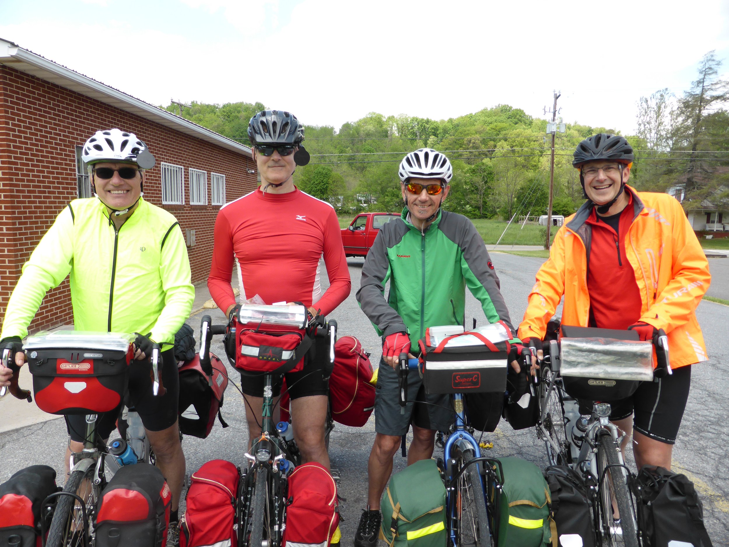 Four smiling men in cycling gear and helmets standing with bikes, outdoors near a brick building, green trees, and power lines.