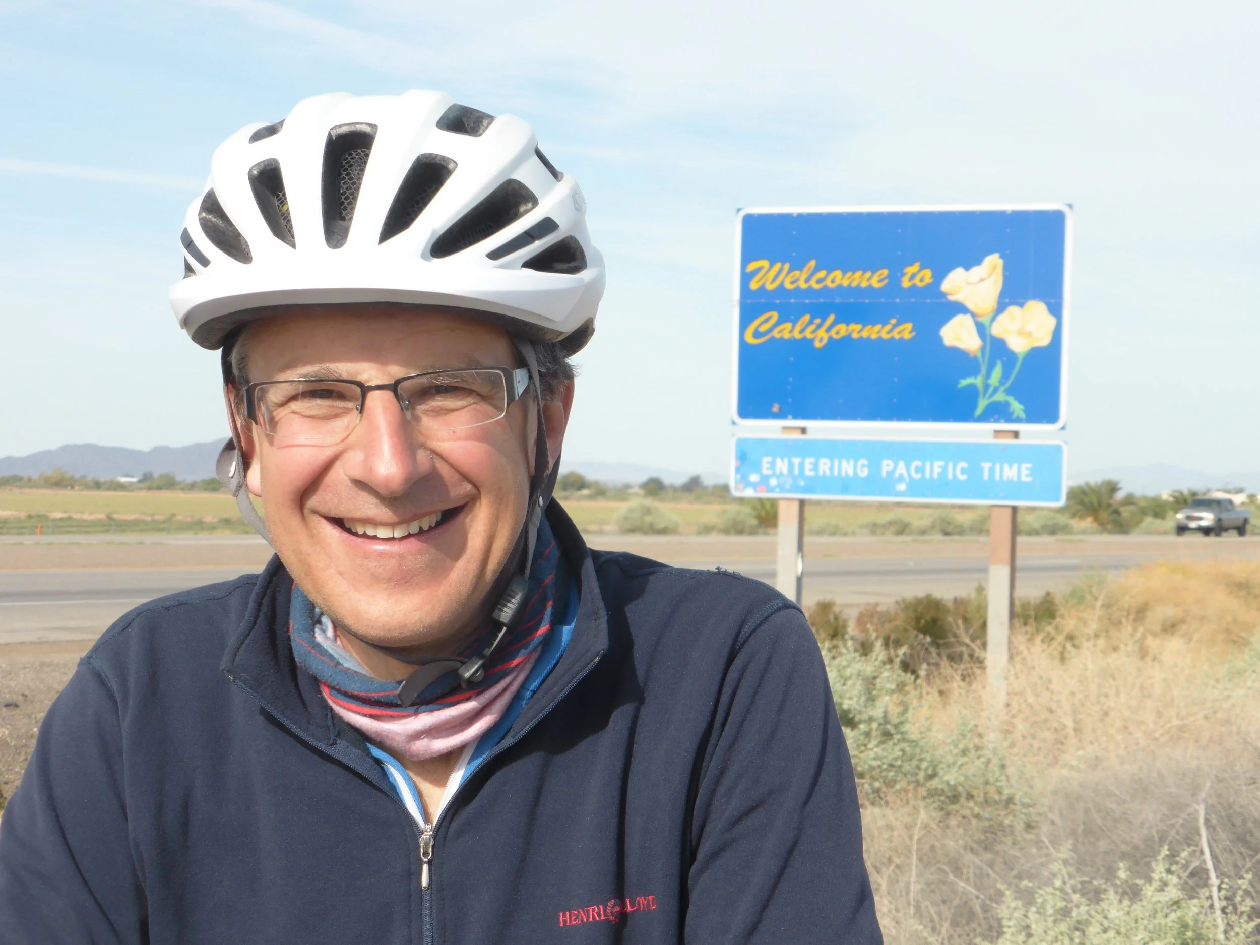 A man wearing glasses, a bicycle helmet, and a navy jacket smiling for a photo in front of a 'Welcome to California' sign on the side of a highway.