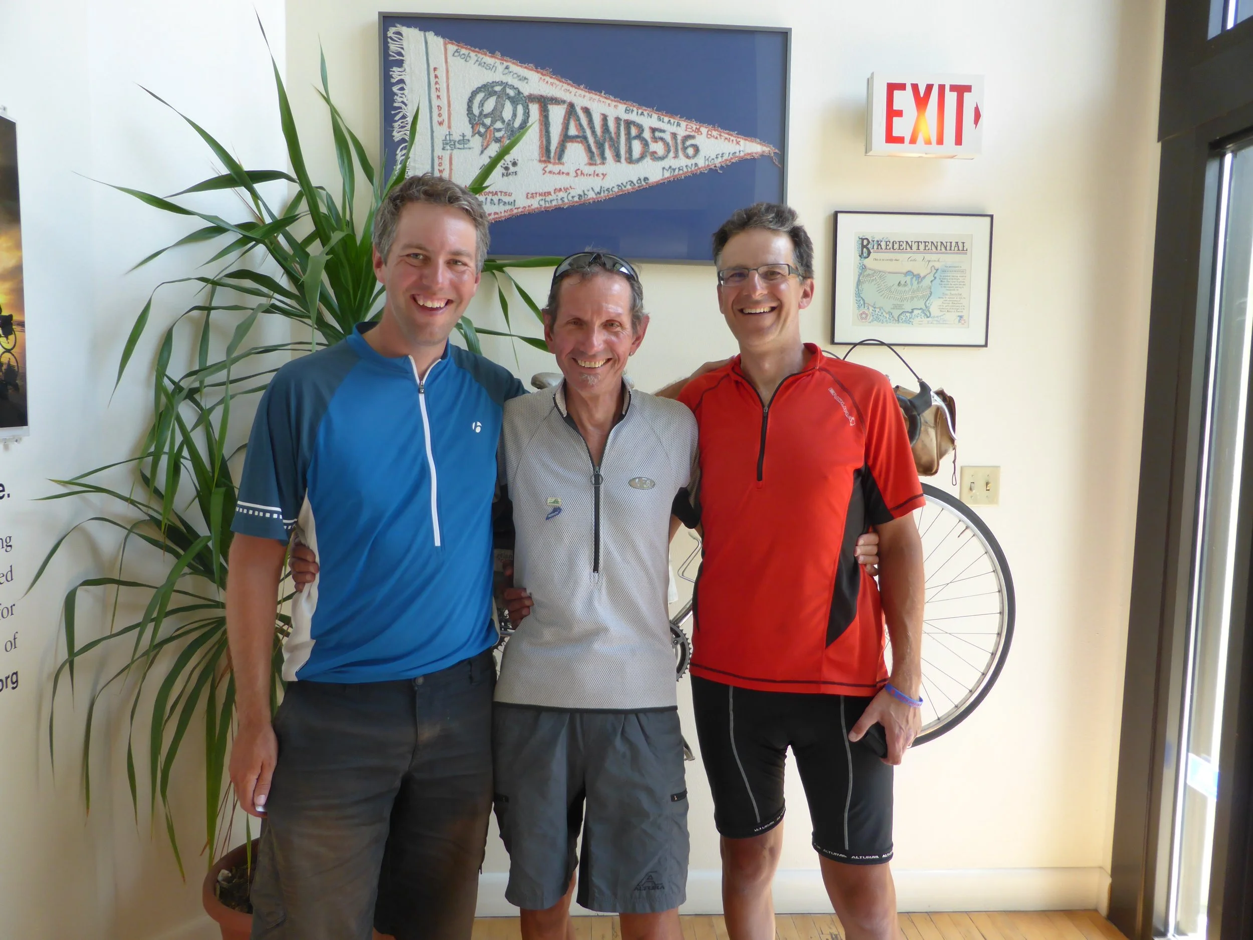 Three men standing together indoors, smiling. The man on the left is wearing a blue cycling jersey, the man in the middle is wearing a gray shirt, and the man on the right is wearing a red cycling jersey. Behind them are framed posters and a large green plant, with a bicycle wheel partially visible on the right.
