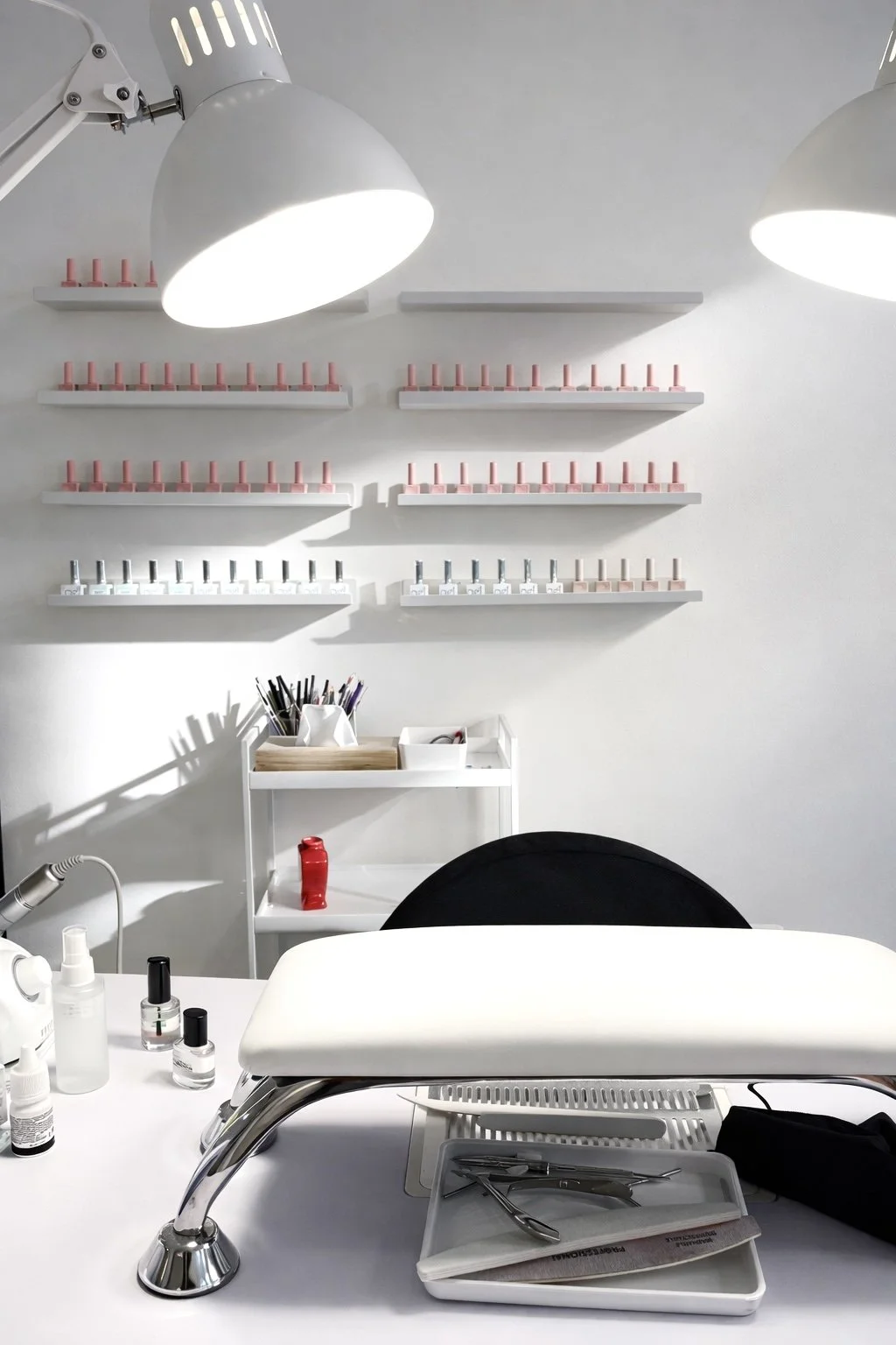 Een schoonmaaktafel in een schoonheidssalon of nagelstudio met gereedschap en polishes, witte muren en verzamelplaatsen voor nagellak. Boven de tafel hangen verschillende rekken met houderkasten voor nagellak.
