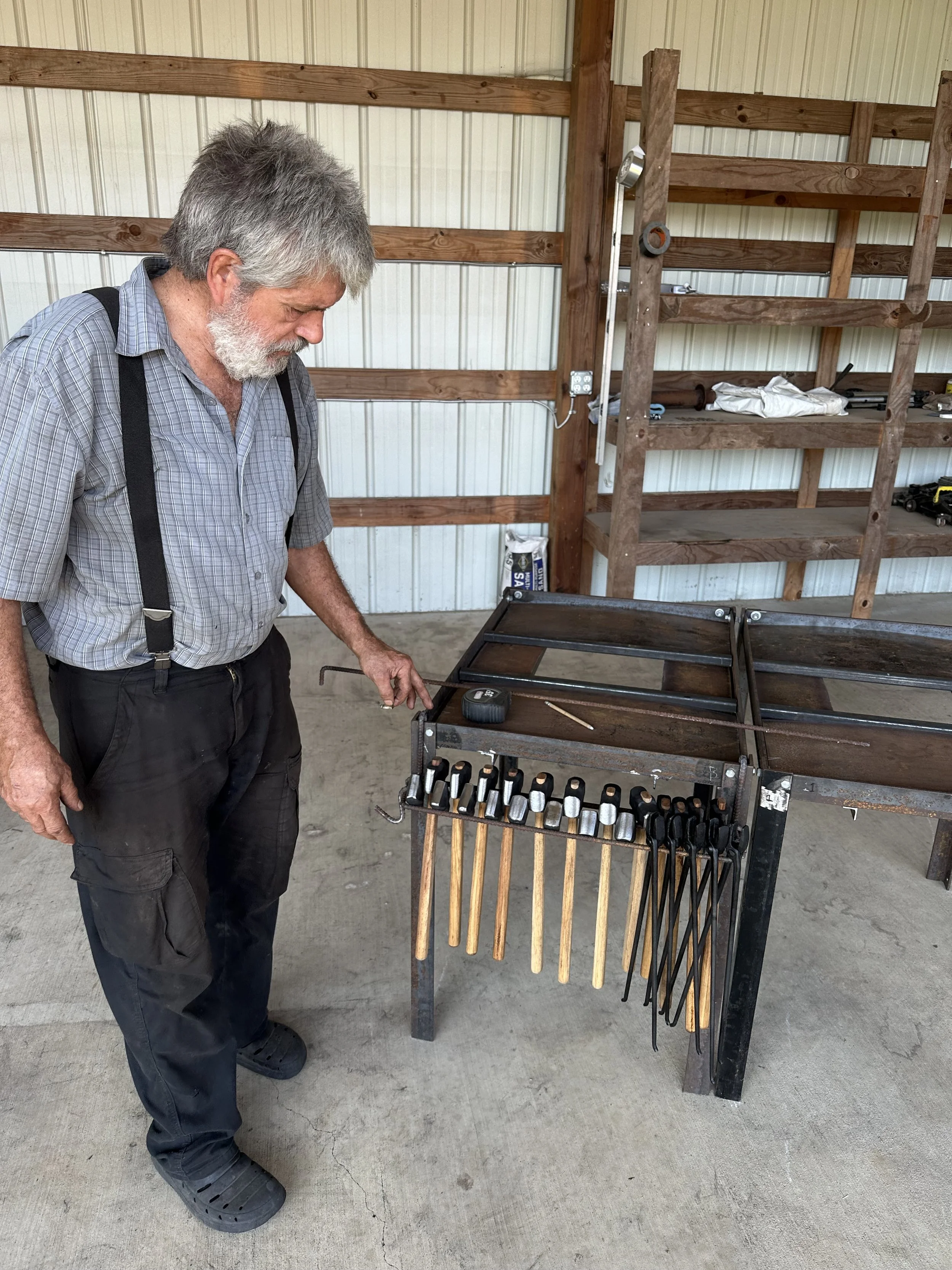An elderly man with gray hair and beard examining a black metal and wooden tool stand with various metal and wooden-handled tools inside a workshop with wooden shelves and tools on the background.