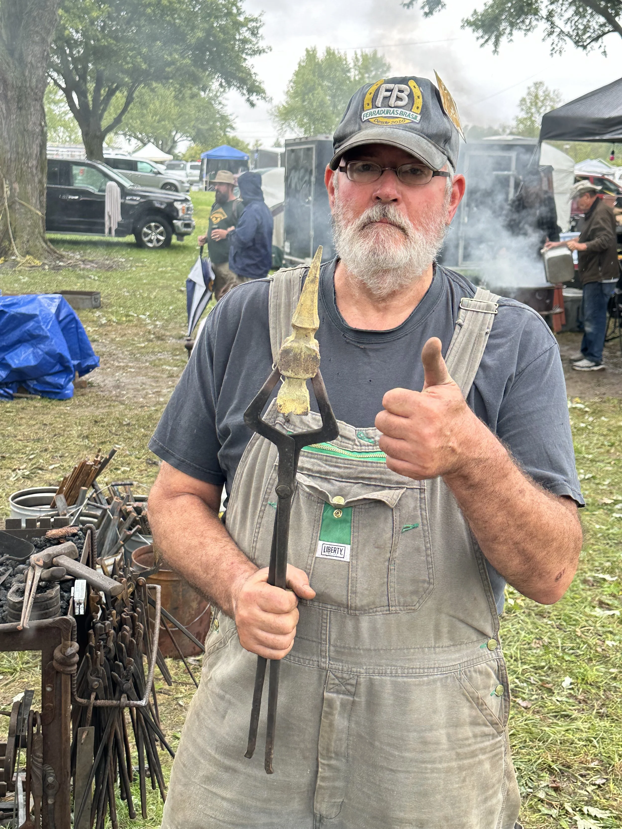 A man with a gray beard, wearing glasses, a gray cap, a dark shirt, and tan overalls, stands outdoors at a blacksmith event, holding a heated metal piece on blacksmithing tongs and giving a thumbs-up.