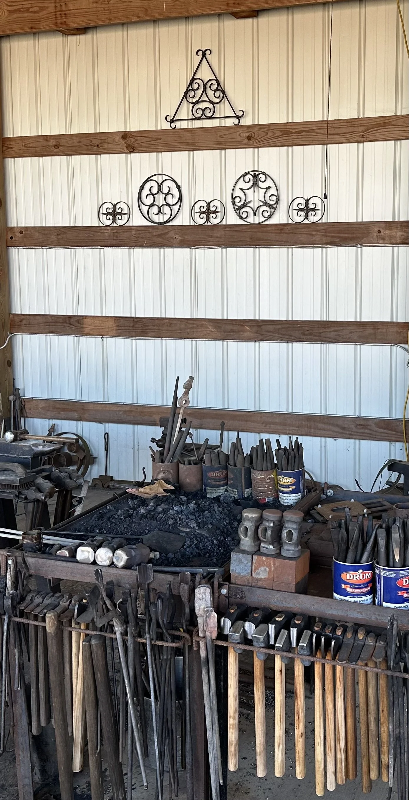 Blacksmith's workshop with tools, hammers, tongs, and metal artifacts on a workbench; decorative wrought iron wall art hanging on a wall with wooden beams.