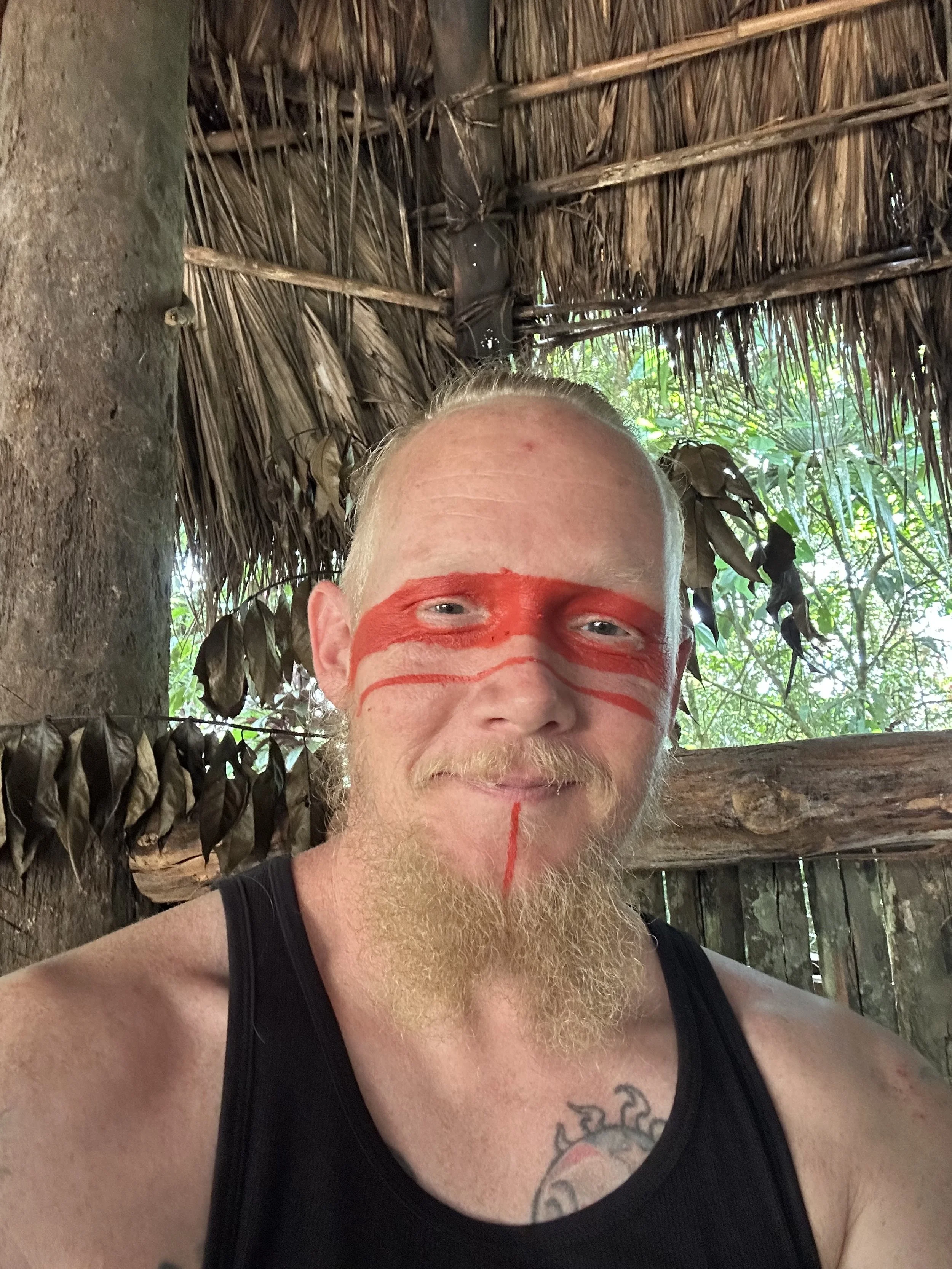 A man with a beard and mustache wearing a black tank top and red face paint in a tribal or ceremonial style, standing inside a wooden hut with thatched roof and green foliage visible outside.