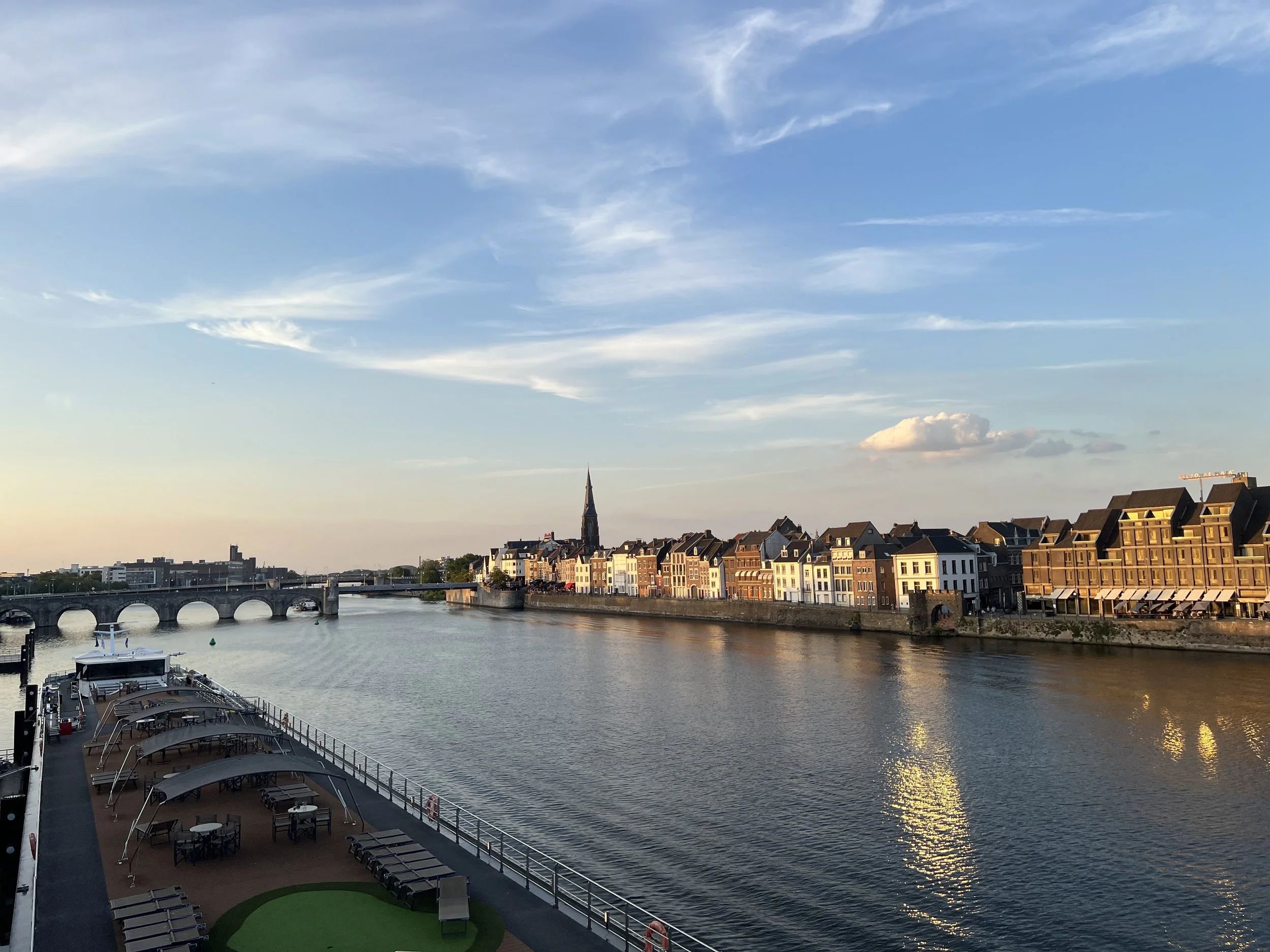 View of a river with reflections, lined by colorful buildings and a church steeple, under a partly cloudy sky during sunset.