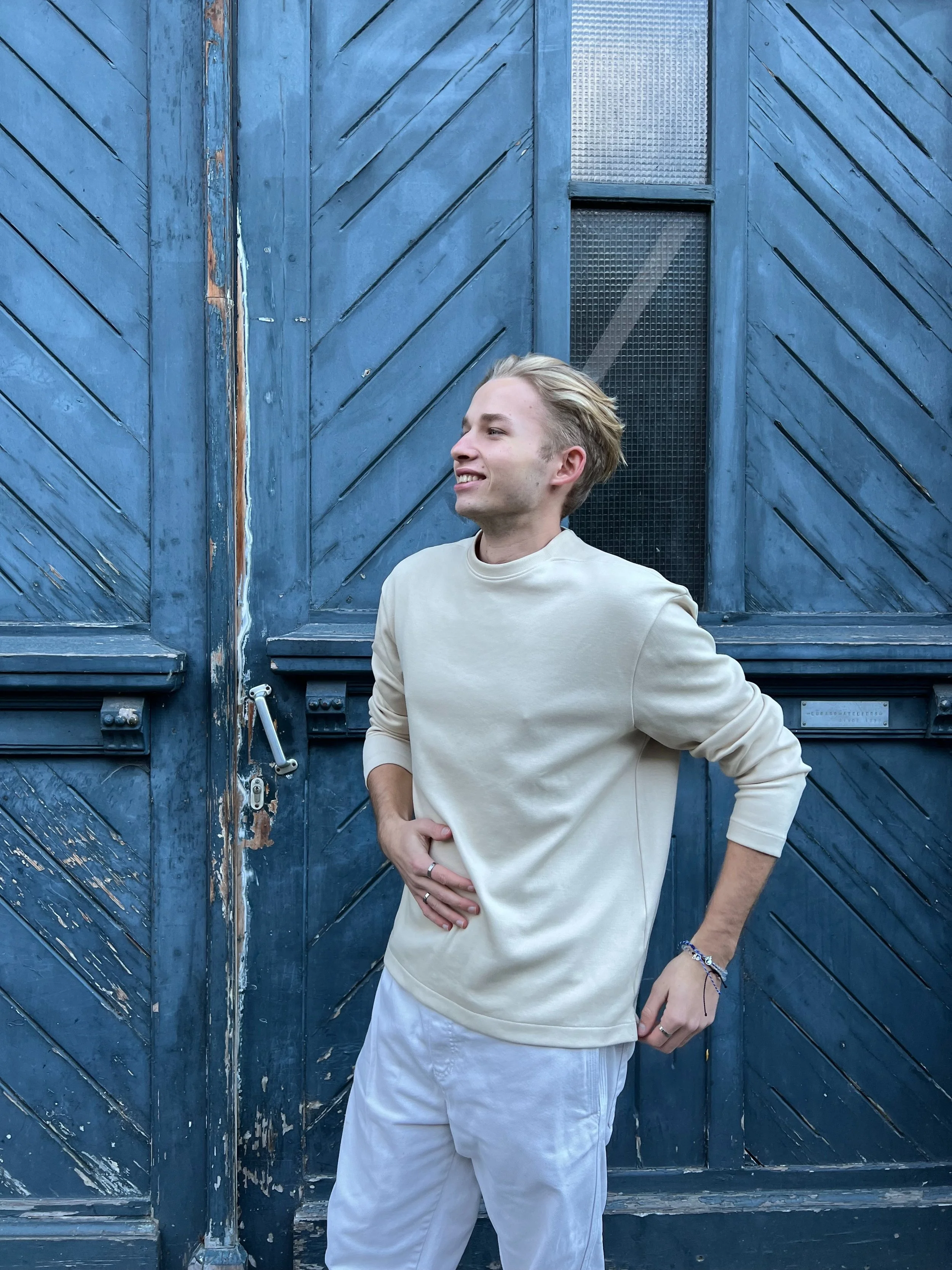 Young man in beige sweatshirt and white pants standing in front of a weathered blue wooden door, smiling and posing.