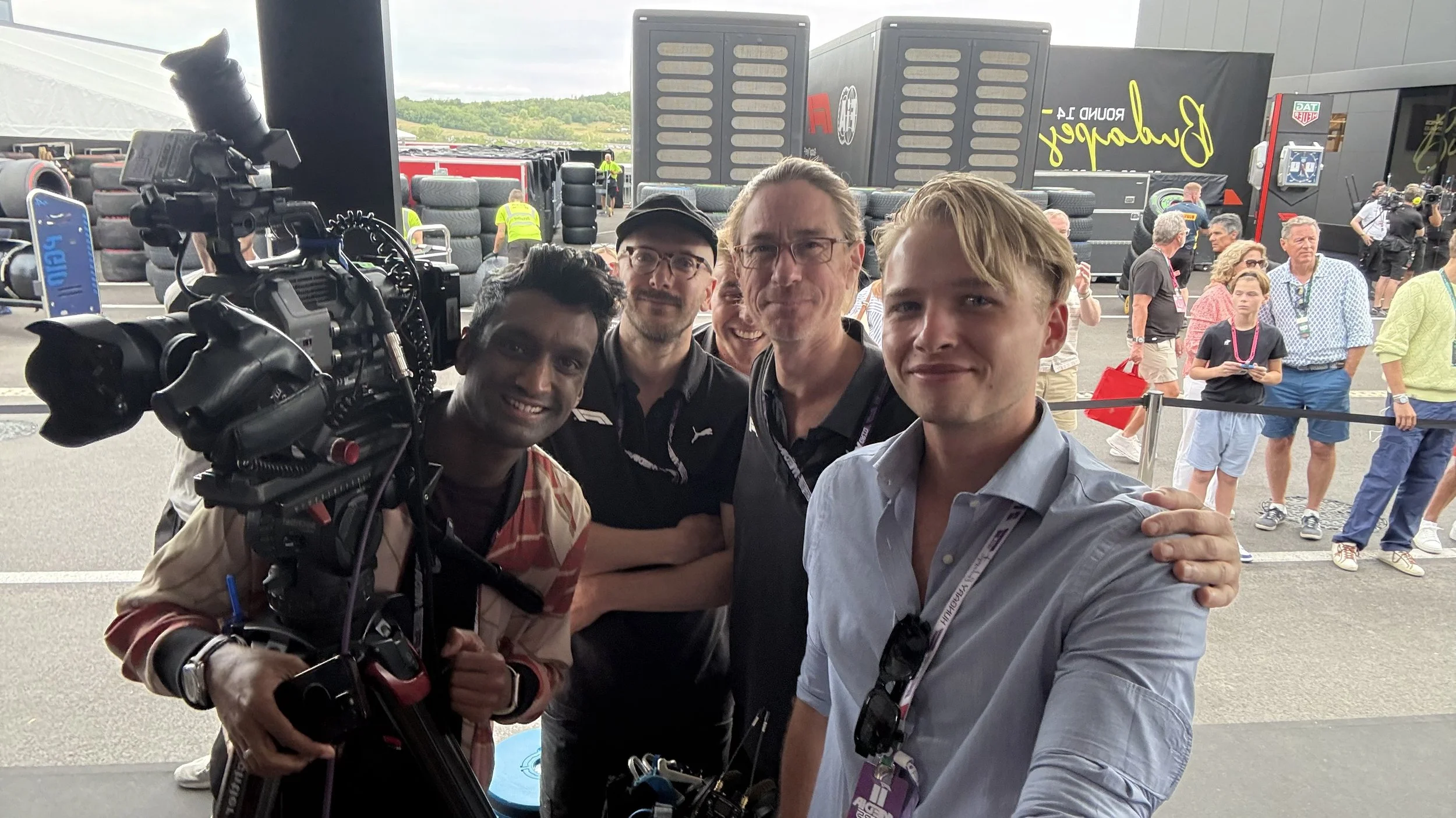 Group of five men smiling and taking a selfie at a racing event with trailers, trucks, and other people in the background.