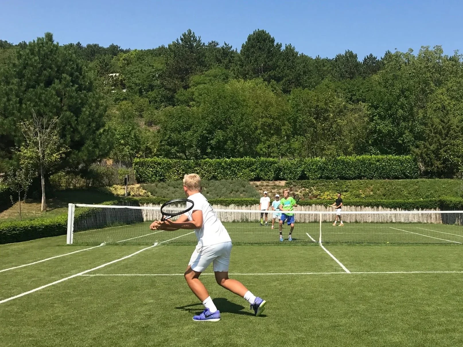 A group of children playing tennis outdoors on a grassy court surrounded by trees.