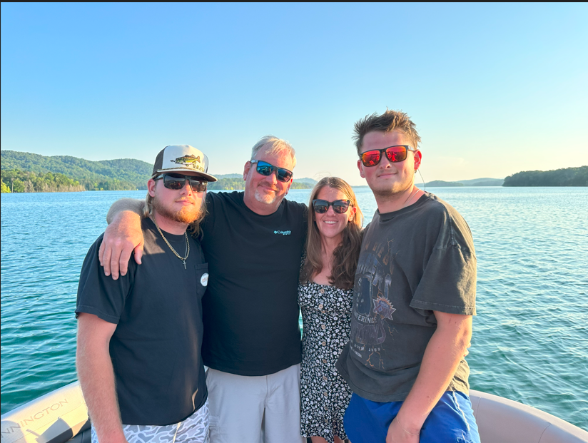 Four people stand on a boat in front of a lake with trees and hills in the background. They are smiling and wearing sunglasses, with one man having his arm around a woman.