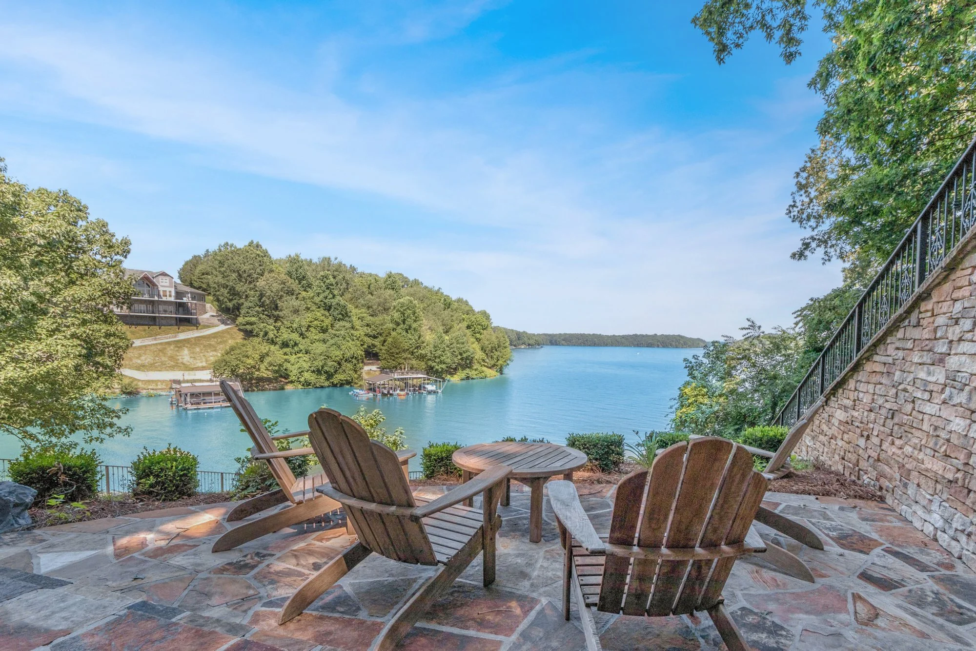 Patio with four wooden chairs and small round table overlooking a lake with trees and houses in the background under a blue sky.