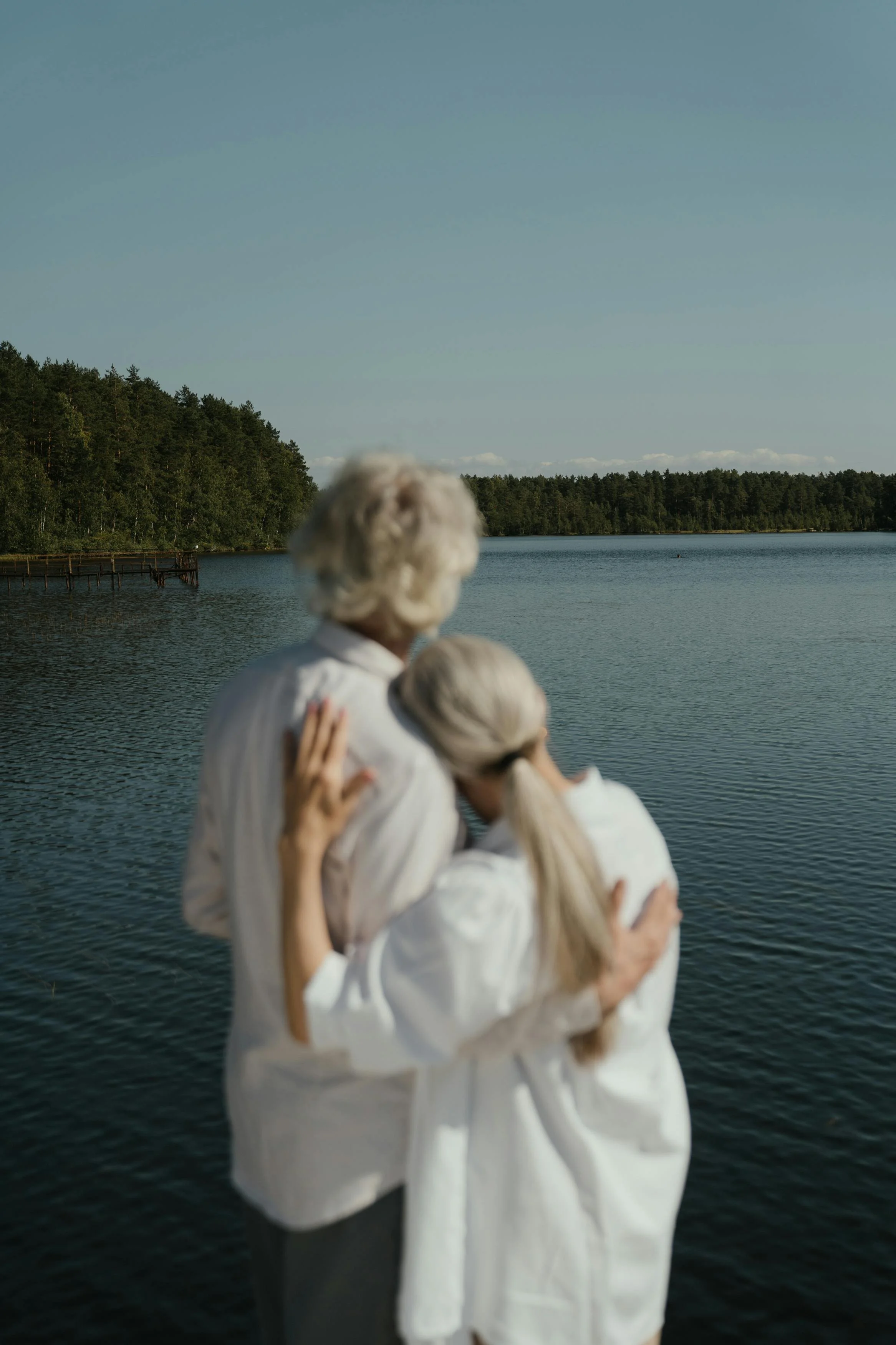A blurred photo of an elderly person with white hair hugging a young girl with long blonde hair, both dressed in white, standing by a lake with a forested shoreline and clear sky in the background.