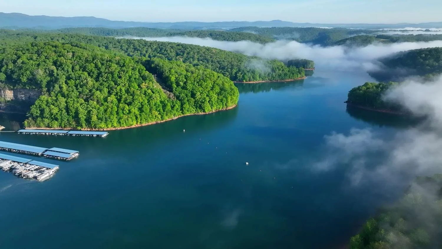 Aerial view of a river surrounded by lush green forested hills with some fog, marina with boats, and distant mountains.