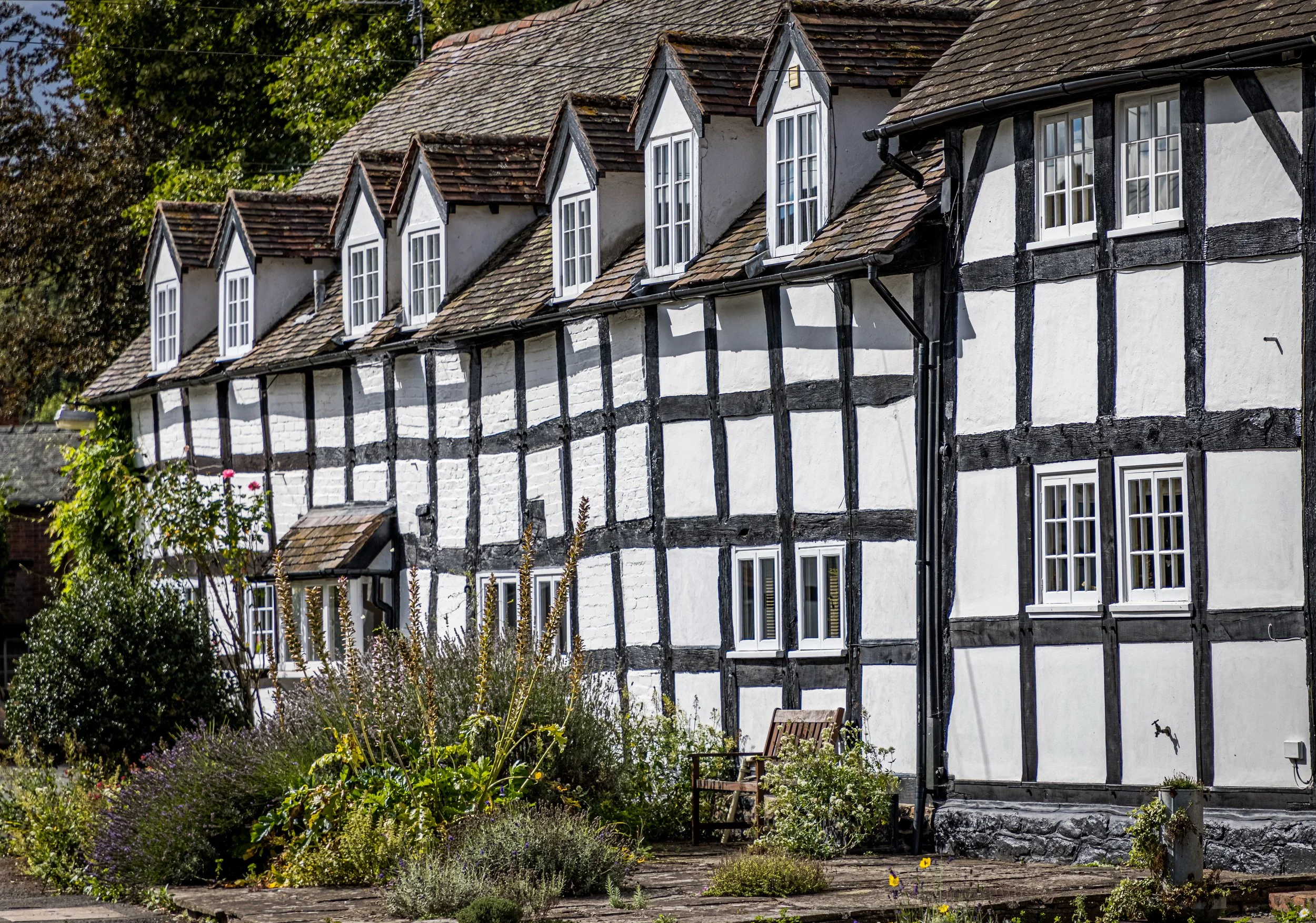 A row of traditional half-timbered houses with black and white facades and sloped roofs, surrounded by greenery and flowering plants.