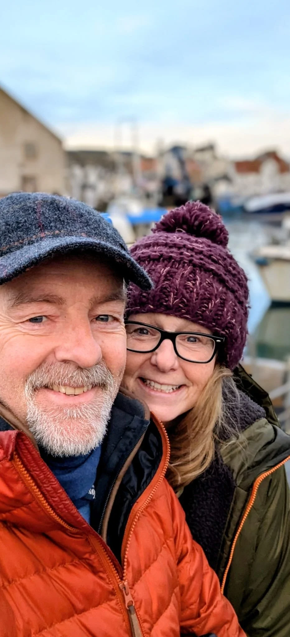 Smiling elderly man and woman taking a selfie at the marina with boats and buildings in the background, wearing warm clothing and hats.
