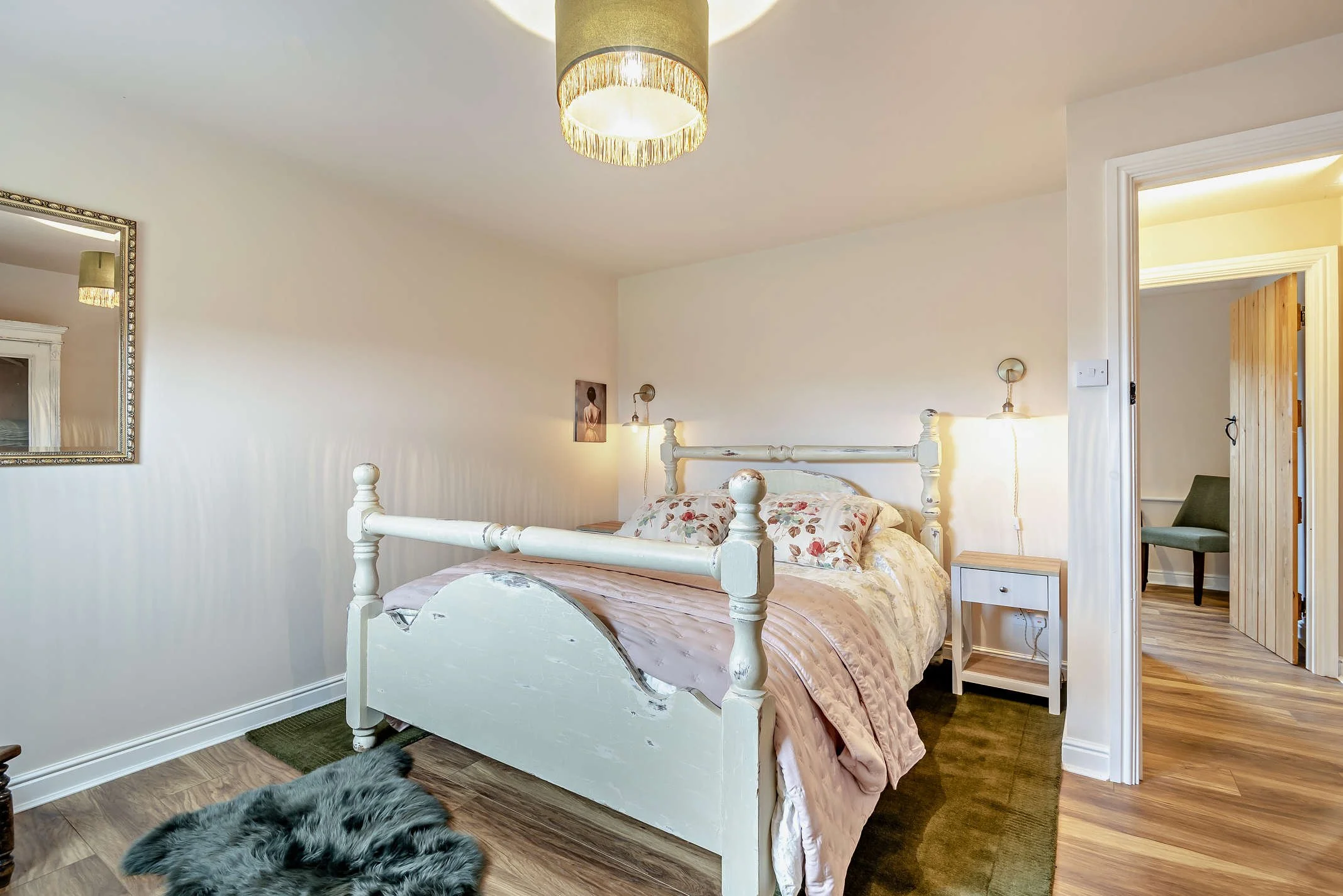 A cozy bedroom with a white vintage-style bed, floral-patterned bedding, a bedside table, and wall-mounted lamps. A large mirror hangs on the left wall, and a small framed artwork is on the wall behind the bed. A doorway shows a glimpse of an adjoining room with a wooden door and a green chair. The room has wooden flooring and soft, warm lighting from a ceiling lamp with fringe.