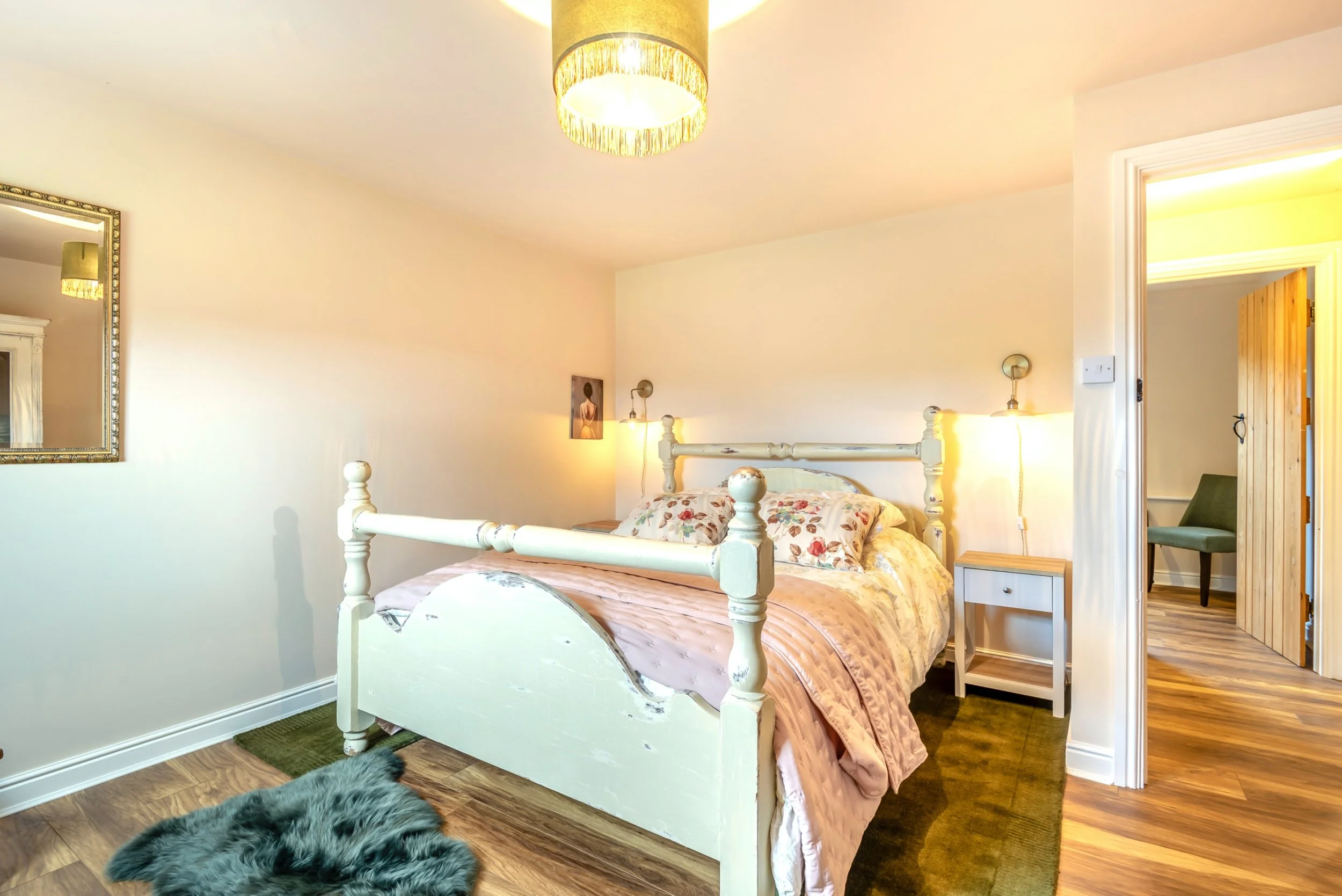 A cozy bedroom with a white wooden bed frame, floral bedding, and pillows. There's a bedside table and lamps mounted on the wall. The room has a mirror on the left wall, artwork above the bed, hardwood flooring, and a furry rug on the floor. An open doorway shows a glimpse of a wooden wall and green chair.