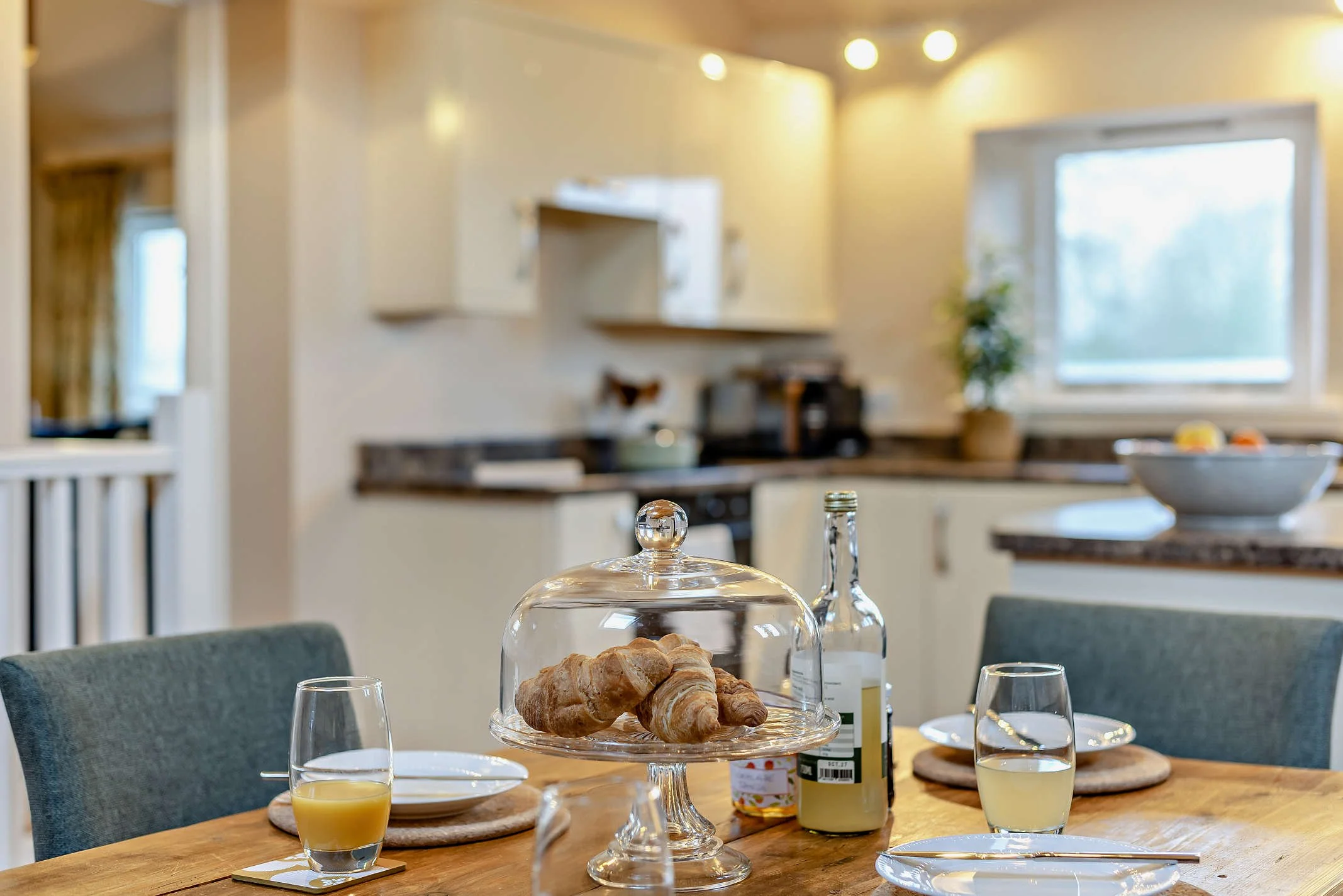 A breakfast table set with a glass dome covering croissants, glasses of orange juice, and bottles of jam or syrup in a cosy kitchen with natural light.