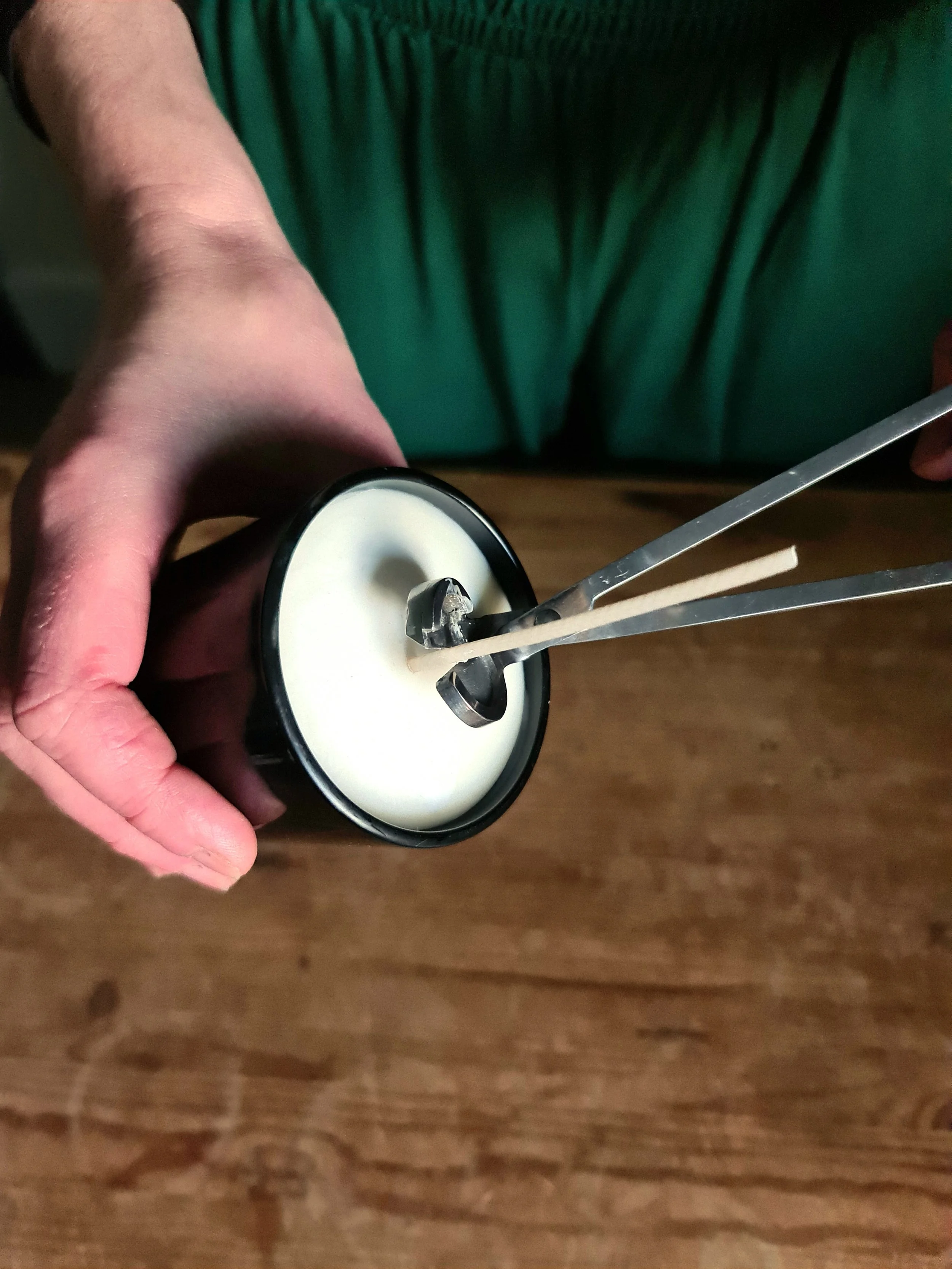 A person holding a black container filled with a white, creamy substance while using a metal tool to stir or scoop it. The person is wearing a green shirt or apron.