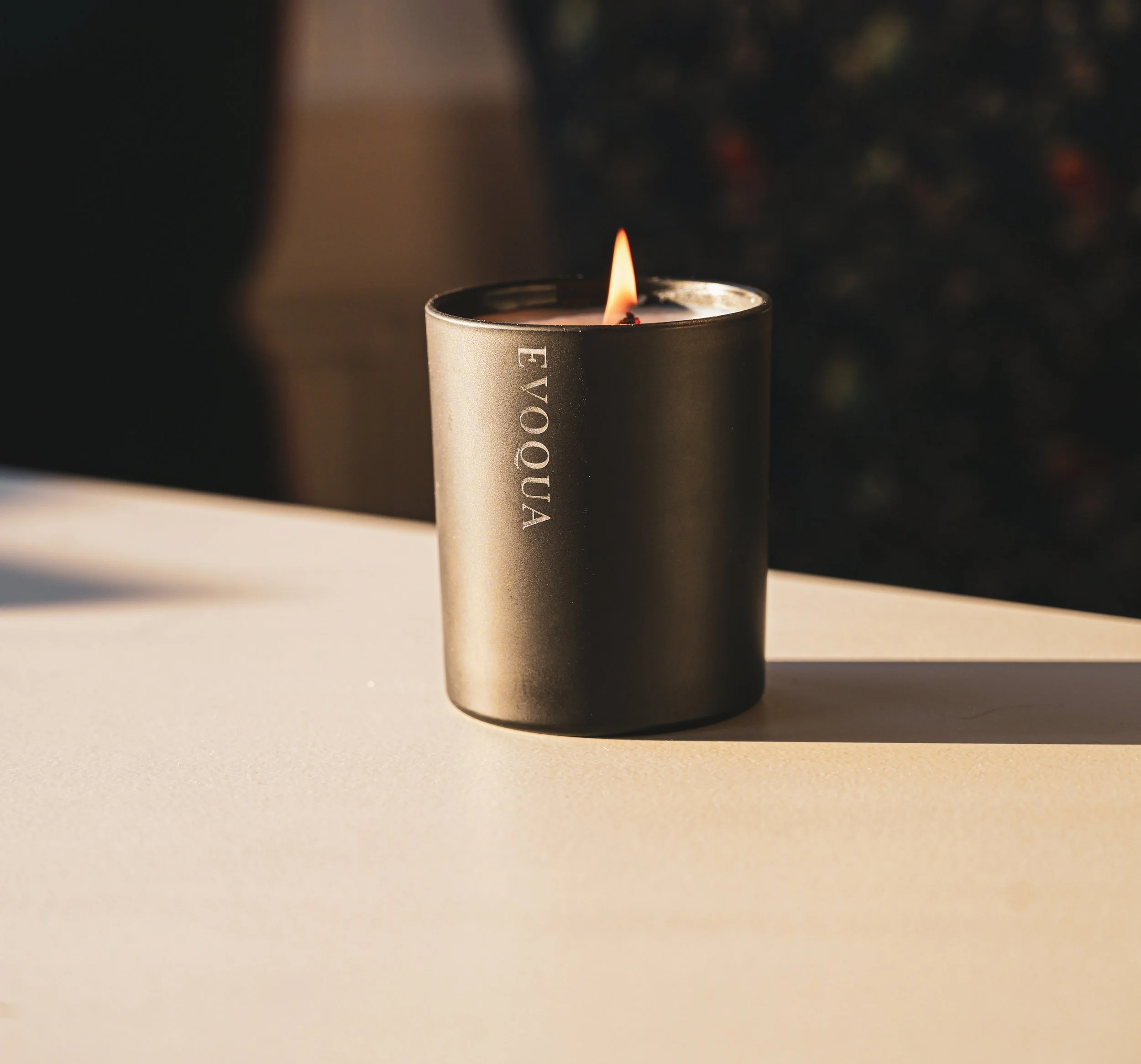 A lit candle in a black holder with the word 'EVOQUA' printed on it, placed on a white surface against a dark, blurred background.