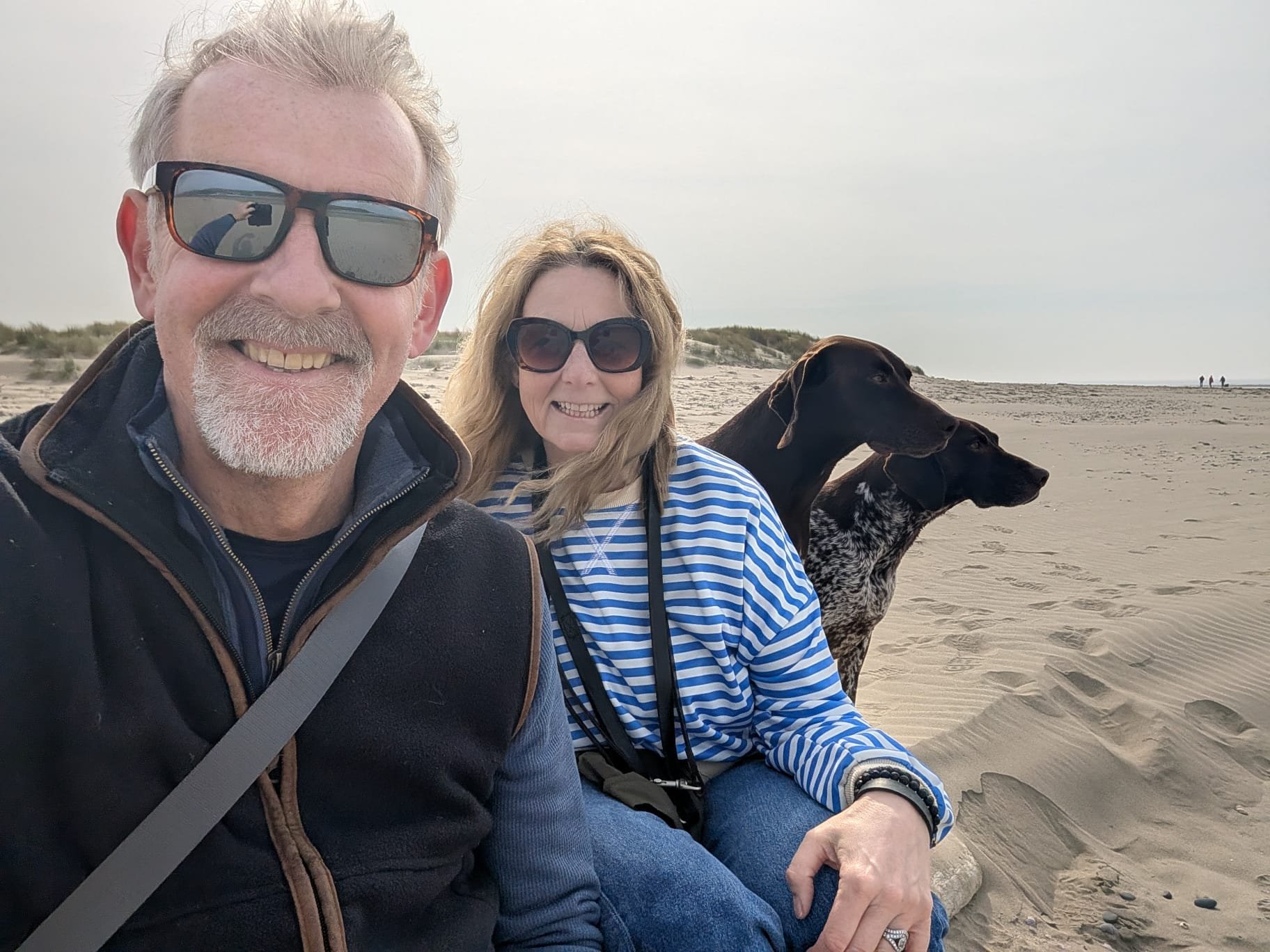 Smiling older man and woman sitting on the sand at the beach, with two dogs, one black and the other black and white, beside them. The man is wearing sunglasses and a black jacket, the woman is wearing large dark glasses and a striped blue and white top. There are sand dunes in the background and a few people walking in the distance.