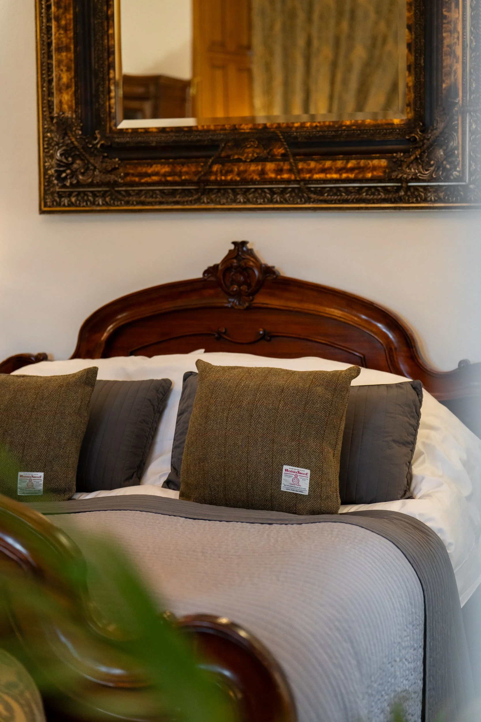 A close-up of a bed with a carved wooden headboard, featuring a large framed mirror on the wall above it. The bed has white bedding, with several decorative throw pillows in shades of brown and gray. The headboard and pillows exhibit a classic, vintage style.