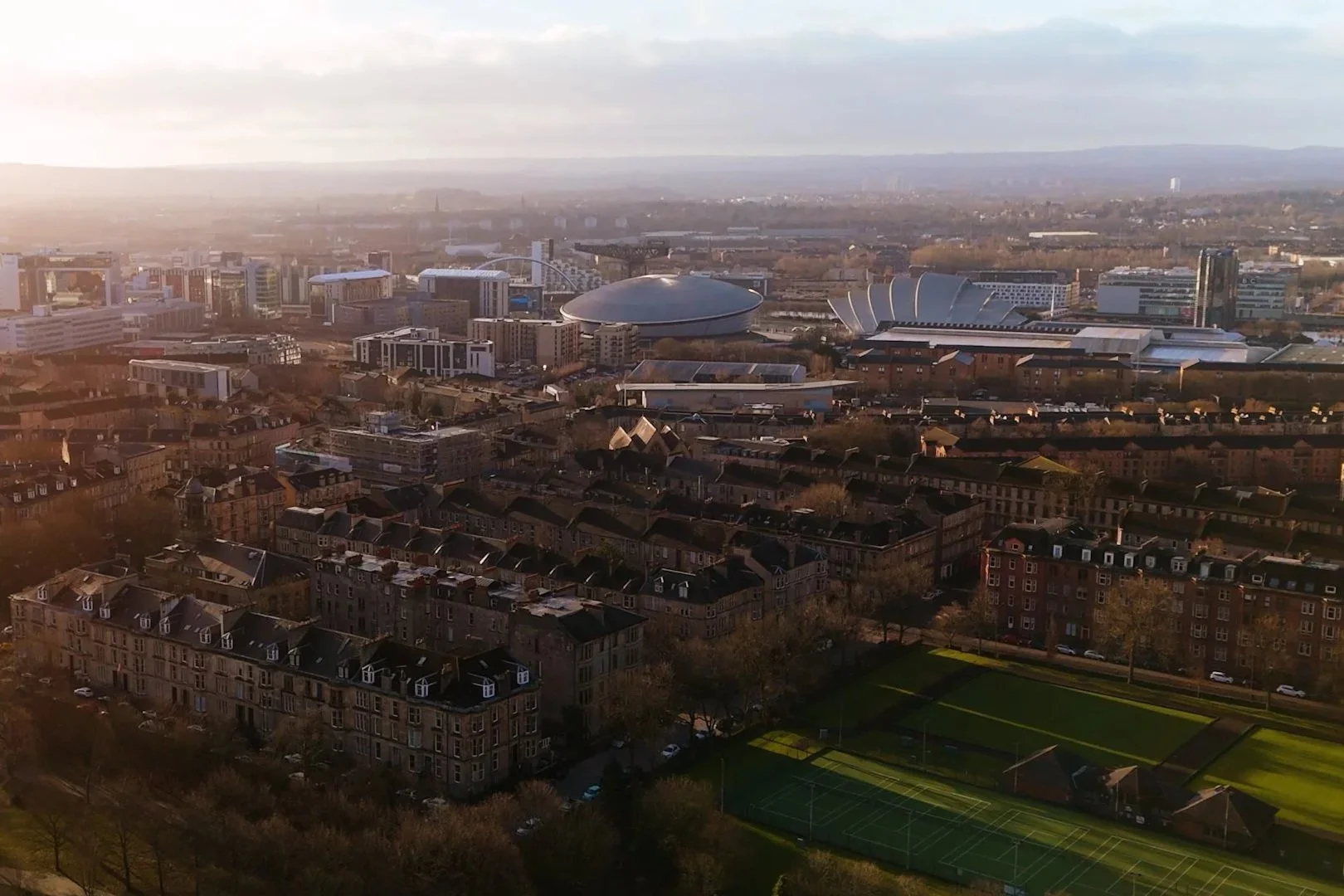 Aerial view of a city skyline with modern and historic buildings, sports fields, and a stadium in the distance, all under a partly cloudy sky during sunset.