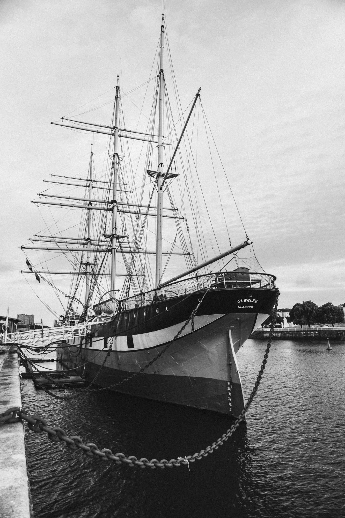 A black and white photo of a large sailing ship docked at a harbor with multiple masts and rigging, named Glenlee, with the cityscape visible in the background.