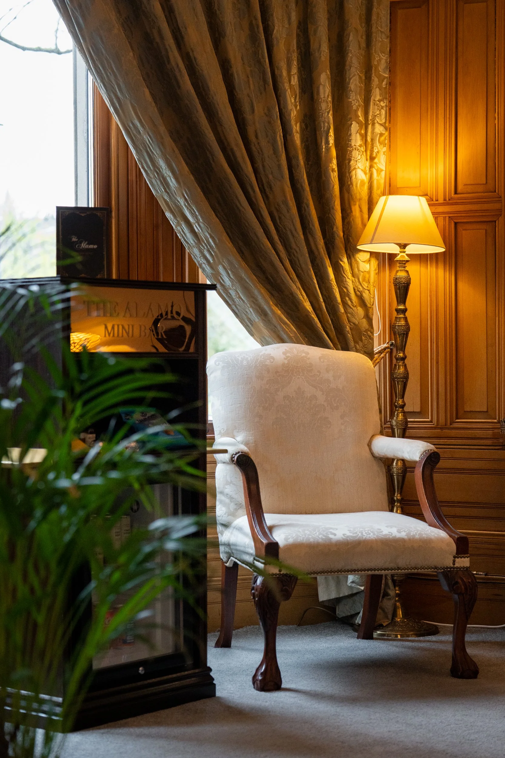 A cozy corner featuring a plush, vintage armchair with wooden carved legs, next to a wooden-paneled wall with a standing lamp, partially covered window with heavy, patterned curtains, and a small mini bar refrigerator.